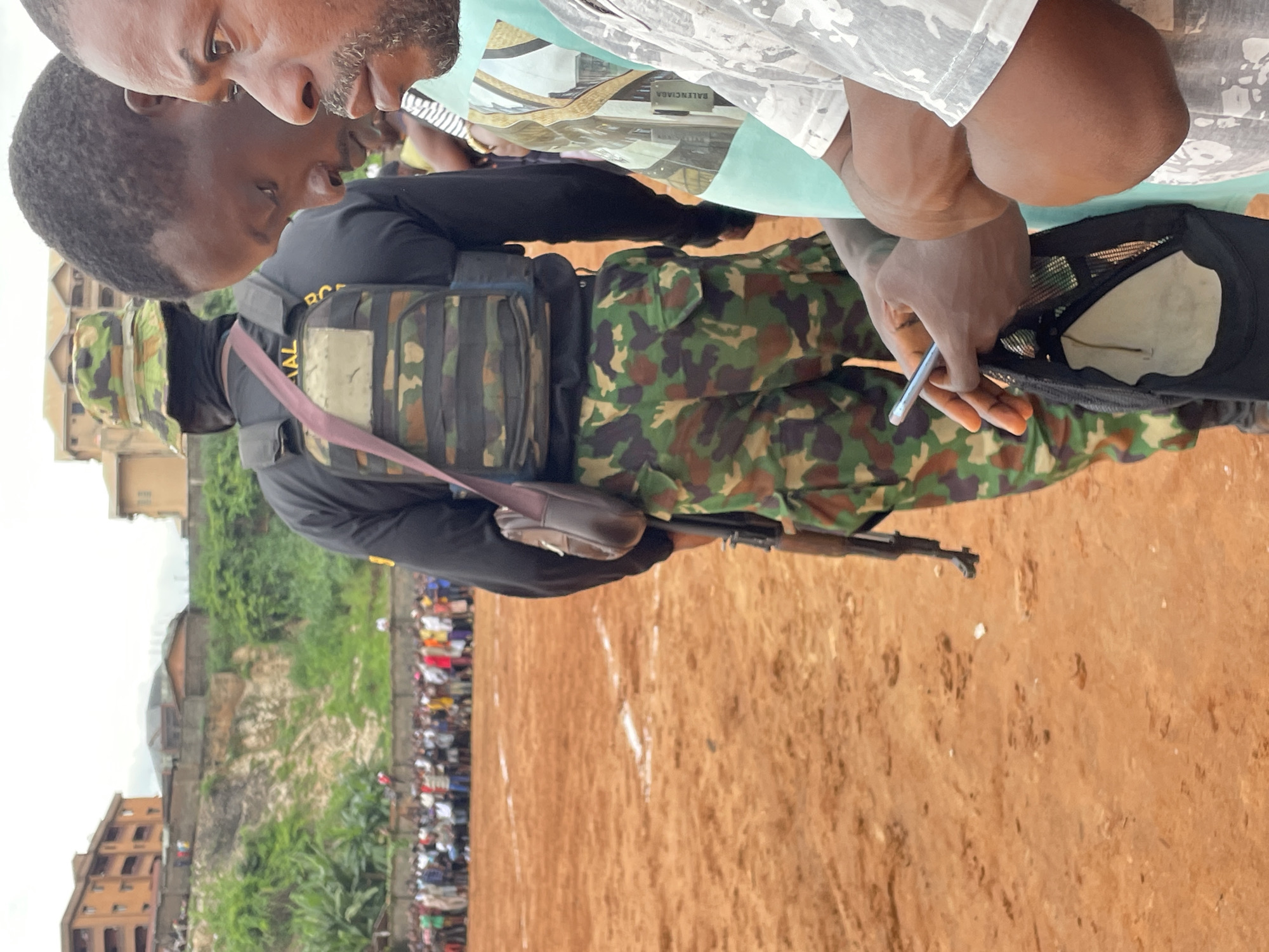In an outdoor scene, the focus is on three men in the foreground, with a crowd of people and buildings in the background. The man on the left, viewed from the back, wears a camouflage hat, a black long-sleeved shirt, a tactical vest with camouflage patterns, and camouflage pants. A brown leather shoulder bag is slung across his chest, and a rifle is partially visible hanging from his shoulder. The man in the middle, viewed in profile, has dark skin and hair, and is looking downwards. The man on the right, also in profile, has darker skin and a beard, and is looking to the left. His right arm is bent, and his left arm is extended, holding a smartphone. He is wearing a light blue t-shirt with a graphic print and a white t-shirt with a camouflage pattern. The ground in the foreground is reddish-brown dirt with some white markings. In the mid-ground, a crowd of people of various ethnicities and ages is gathered. Behind the crowd, there are buildings with orange and brown facades, and a hillside covered with greenery. The sky is overcast.