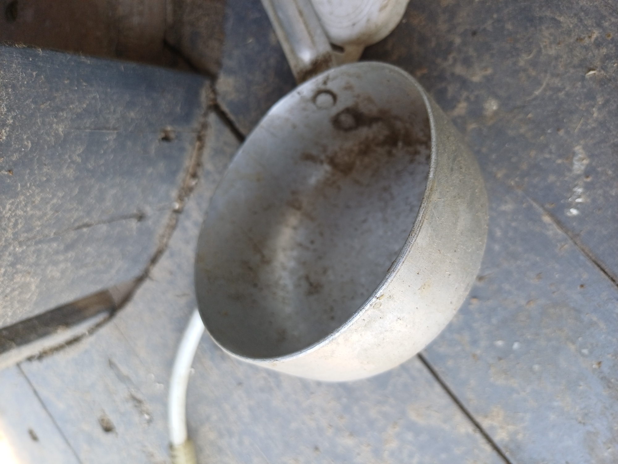 The image shows a weathered metal ladle resting on a dark, textured surface that appears to be aged wood or concrete, possibly in an outdoor setting in Sulymivka, Ukraine. The ladle is empty and shows signs of use, with dirt and residue on its interior and exterior surfaces. A white hose or tube is visible in the background, partially obscured. The overall environment suggests an area that is not actively maintained, with a general appearance of disuse or storage.