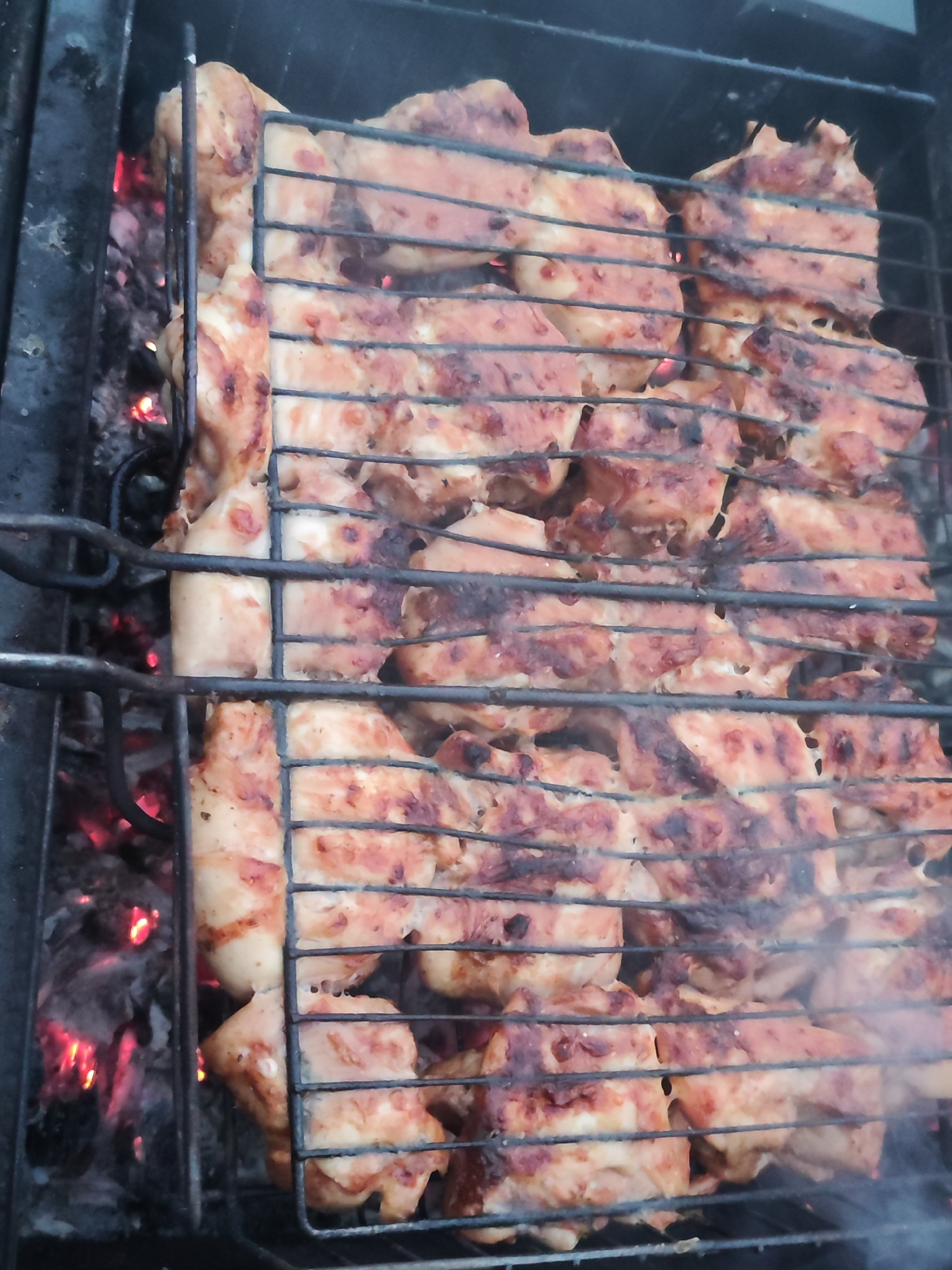 A close-up shot shows marinated chicken pieces grilling on a metal grid over hot coals. The chicken is partially browned with visible grill marks. Some smoke rises from the food, indicating ongoing cooking. The coals underneath are glowing red. The location is unknown and cannot be inferred as Chaikovsky, Russia.