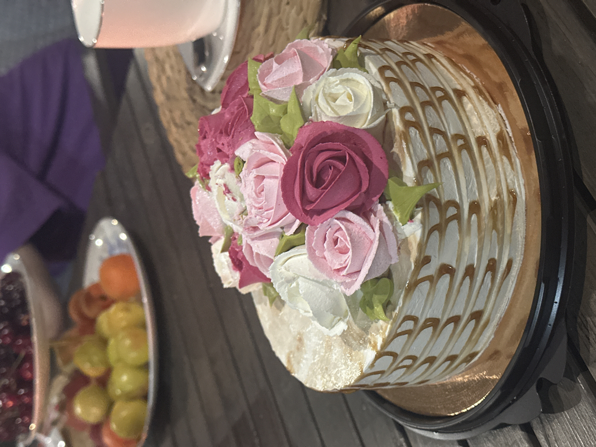 A decorated cake is the central focus of the image. It features white icing with brown caramel drizzle in a lined pattern around the sides, and a cluster of pink, white, and deep pink buttercream roses on top with green leaves. The cake sits on a gold-colored base within a black plastic container. To the left are blurred images of fruits in serving dishes. A purple-clad person is partially visible, also on the left. A wicker-like object is partially visible behind the cake. The image gives no indication of a location in Aviator, Russia.
