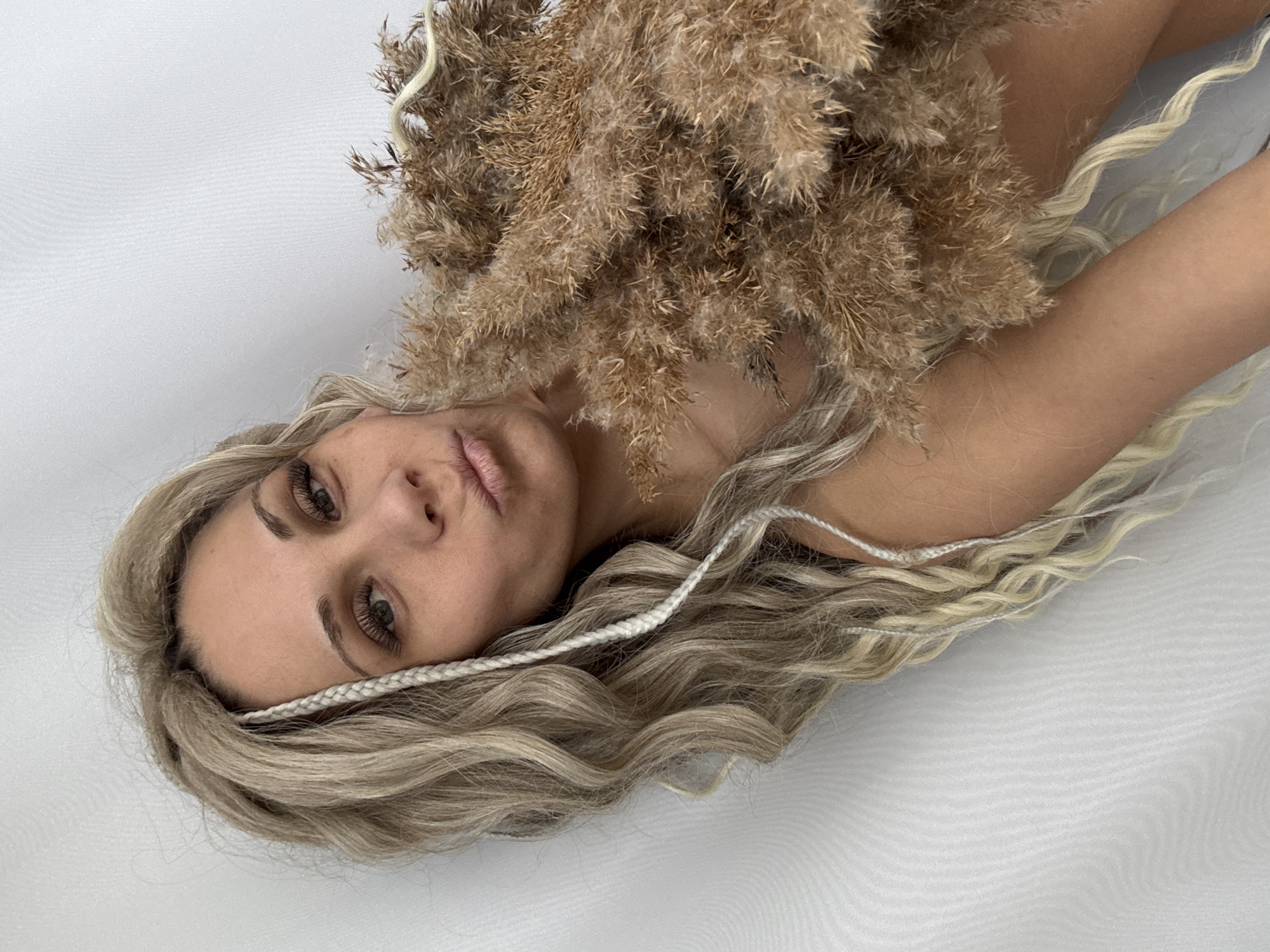 A woman lies on a white surface, her blonde wavy hair spread around her. A bundle of dried pampas grass rests on her chest and shoulder. The woman has fair skin, light eyebrows and eyelashes, and her lips are slightly pursed. A thin braid is visible in her hair. The location cannot be determined from the image.