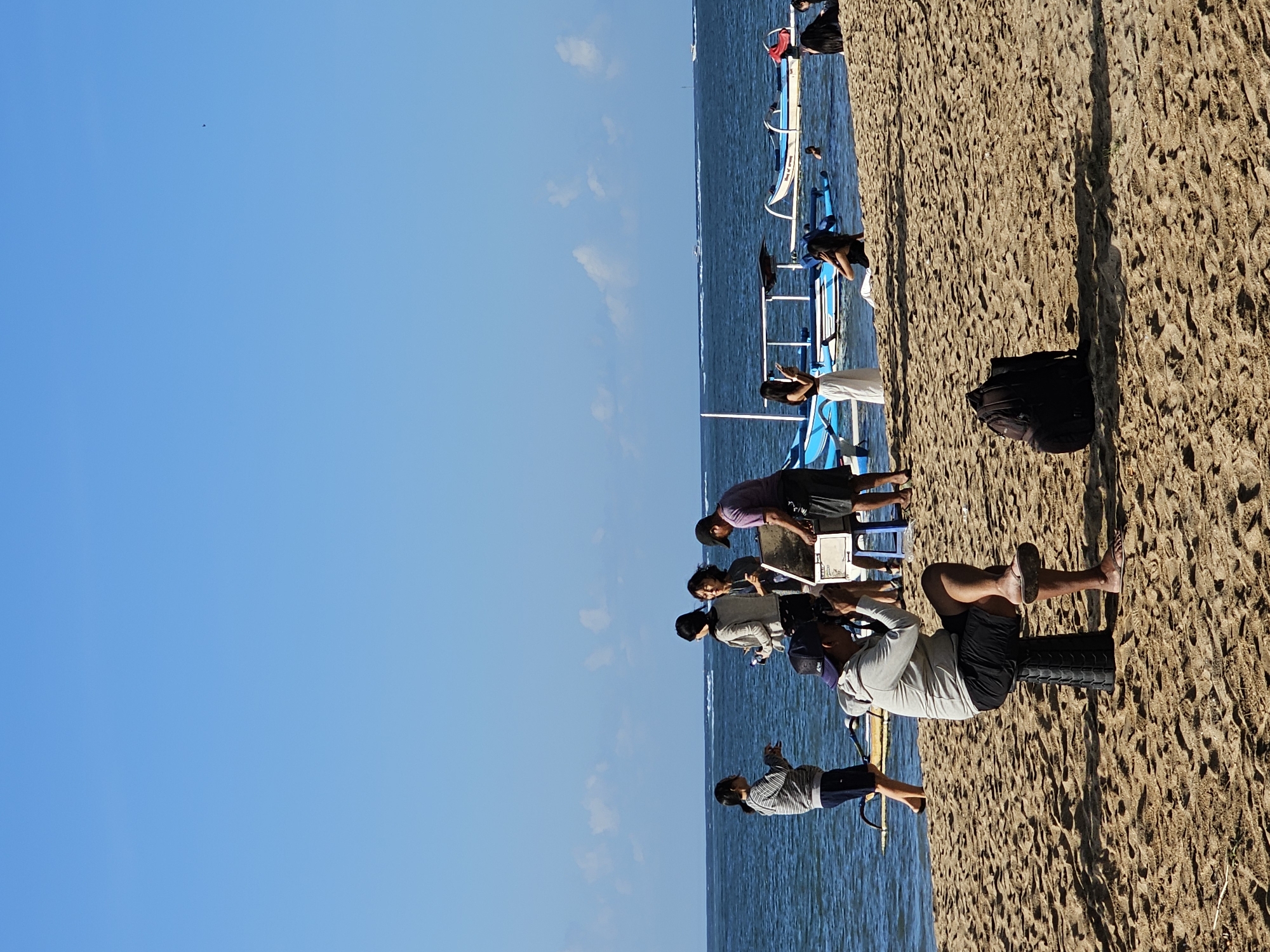 A group of people are on the beach with the ocean and sky in the background. In the foreground, a man is sitting on a black stool and holding a camera up to his face, as if taking a photo or video. He is wearing a grey hooded sweatshirt and black shorts. A black backpack is lying on the sand to his right. In the middle ground, several people are standing near the water's edge. Two women are looking at a display set up by two men. To the right, a man in a purple shirt and shorts is looking at the display, and a woman in a white dress is standing near some blue boats. More blue boats are in the water. In the background, the ocean extends to the horizon under a clear blue sky with a few wispy clouds. The scene appears to be on Kuta Beach in Denpasar, Indonesia, a popular tourist destination known for its sandy shores and recreational activities.