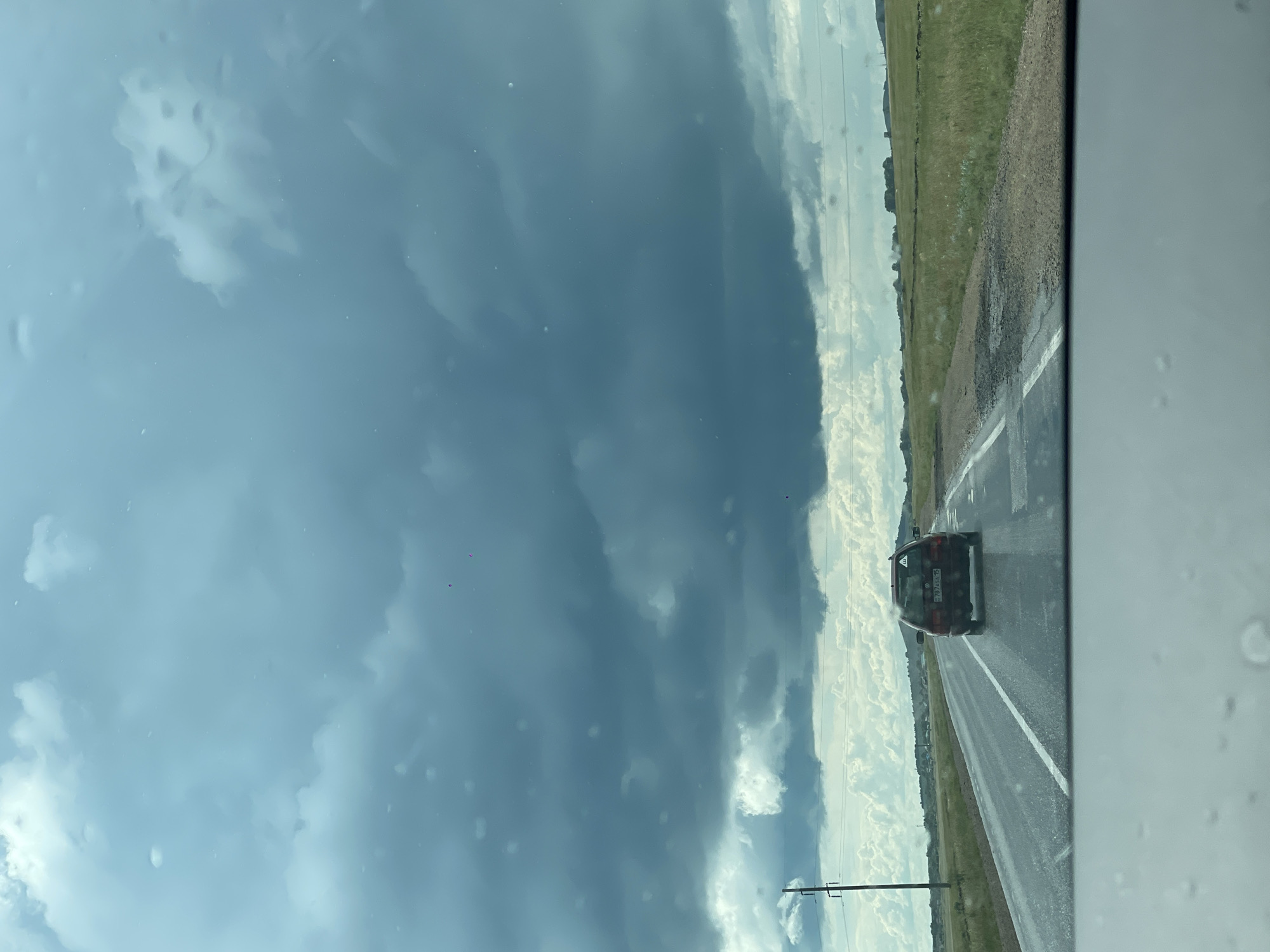A dark, overcast sky with heavy, bruised-looking clouds dominates the upper portion of the image. Light rain or mist is falling, evidenced by water droplets on the windshield in the foreground and the wet appearance of the asphalt road. A red station wagon, with license plate 787 A36 15, is driving away from the viewer on the right lane of the road. The road is flanked by grassy fields. In the distance, a line of trees and some low hills are visible, with power lines and a utility pole to the left of the road. The scene appears to be a rural setting in Kazakhstan, possibly near the city of Жаңаауыл.