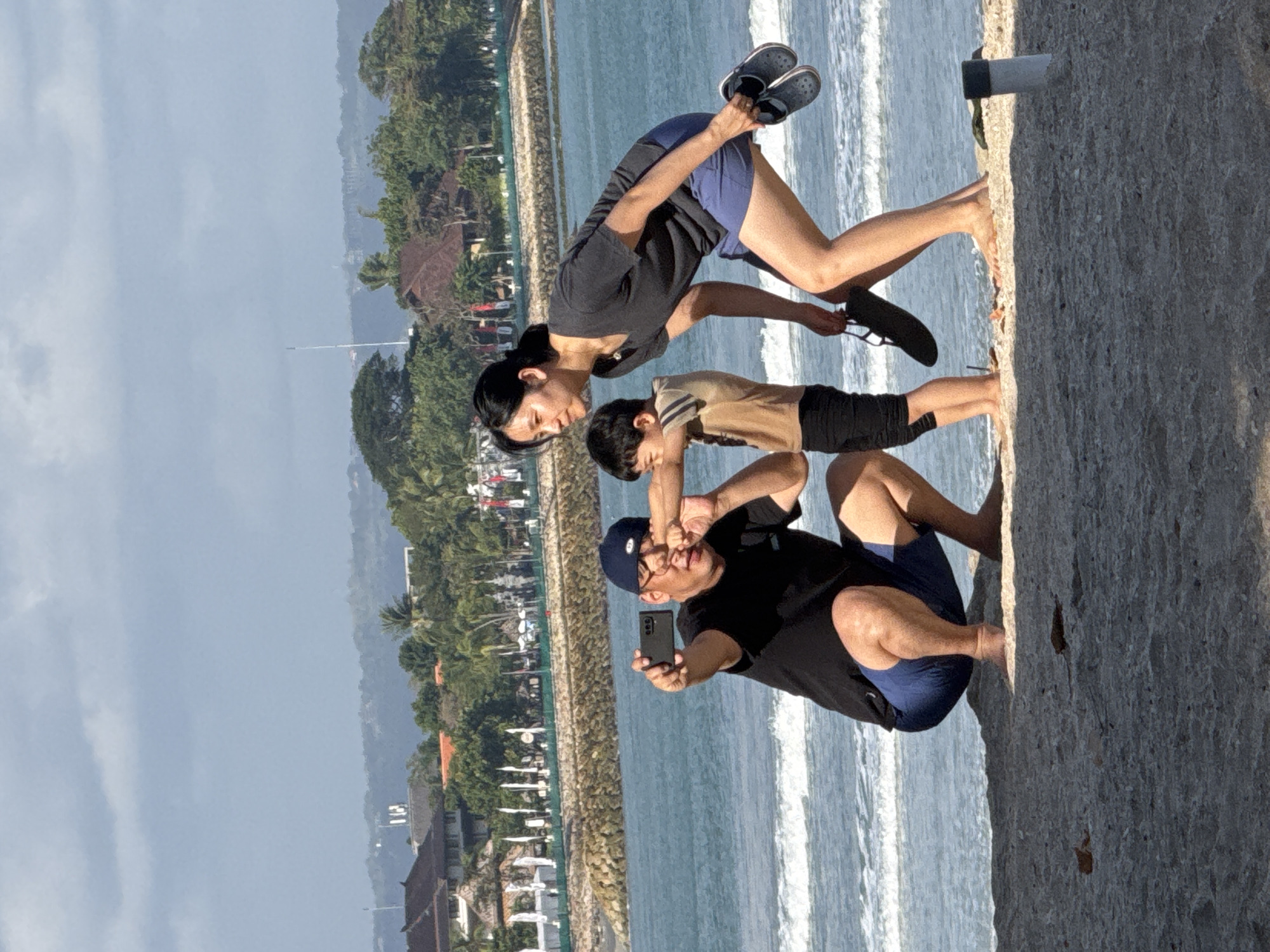 A family is taking a selfie on a beach in Kuta, Indonesia. A man in a black t-shirt and blue shorts is kneeling on the sand, holding up a smartphone to take a picture. A woman in a gray t-shirt and blue shorts is standing next to him, holding a pair of dark-colored clogs in her right hand. A young boy in a tan shirt and black shorts stands between them. The ocean is visible in the background, with waves breaking on the shore. The beach appears to be made of sand and small shells. In the distance, buildings and palm trees can be seen. The sky is partly cloudy.
