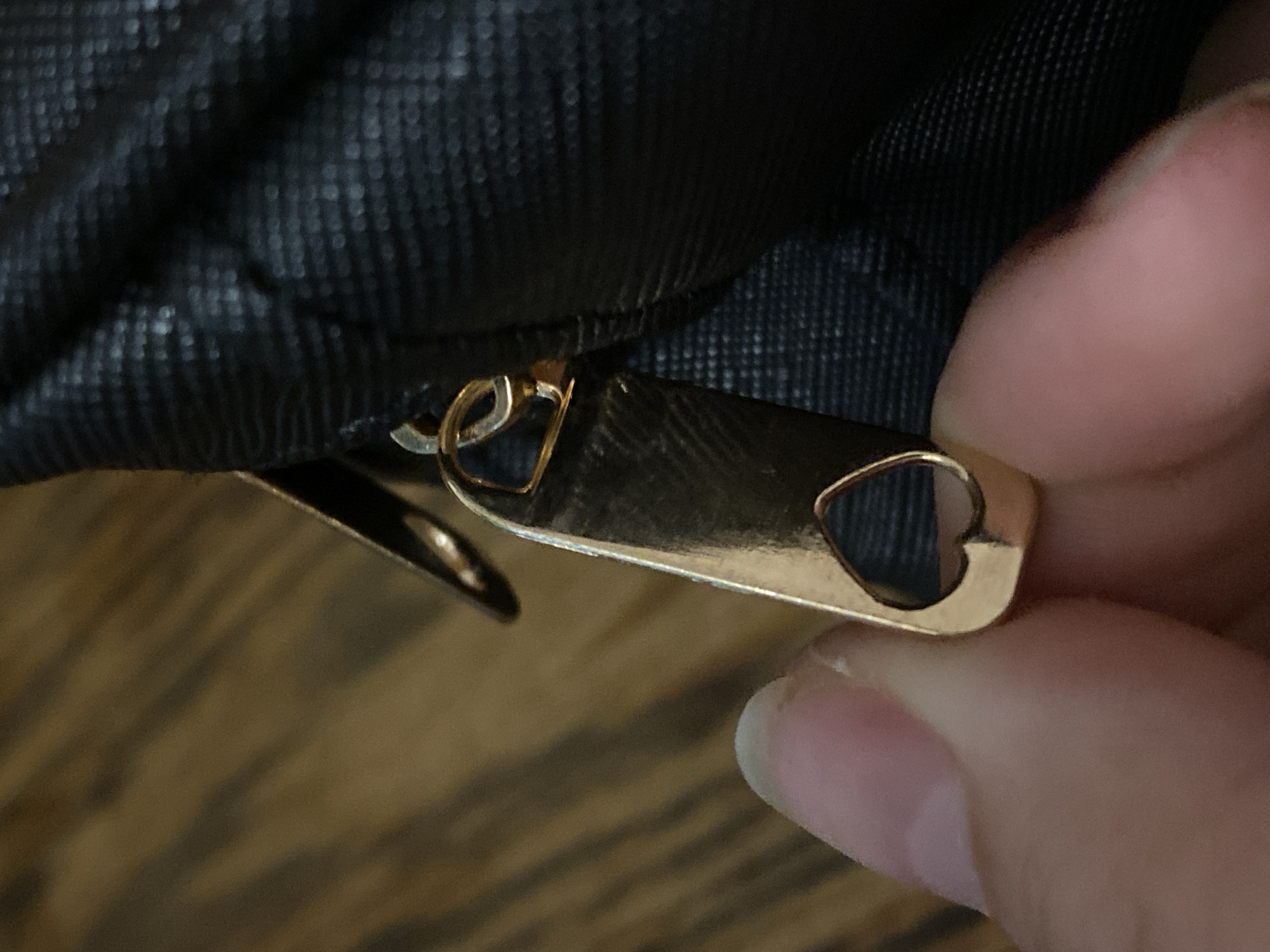 The image shows a close-up view of a gold-colored zipper pull on a black textured fabric, likely part of a bag or garment. The zipper pull is shaped like a heart with a cutout in the center. A finger and thumb are holding the zipper pull, suggesting someone is interacting with it. The background is a blurred wooden surface. The lighting appears to be indoor, with no strong directional shadows, and the colors are warm. There is no visible text in the image.