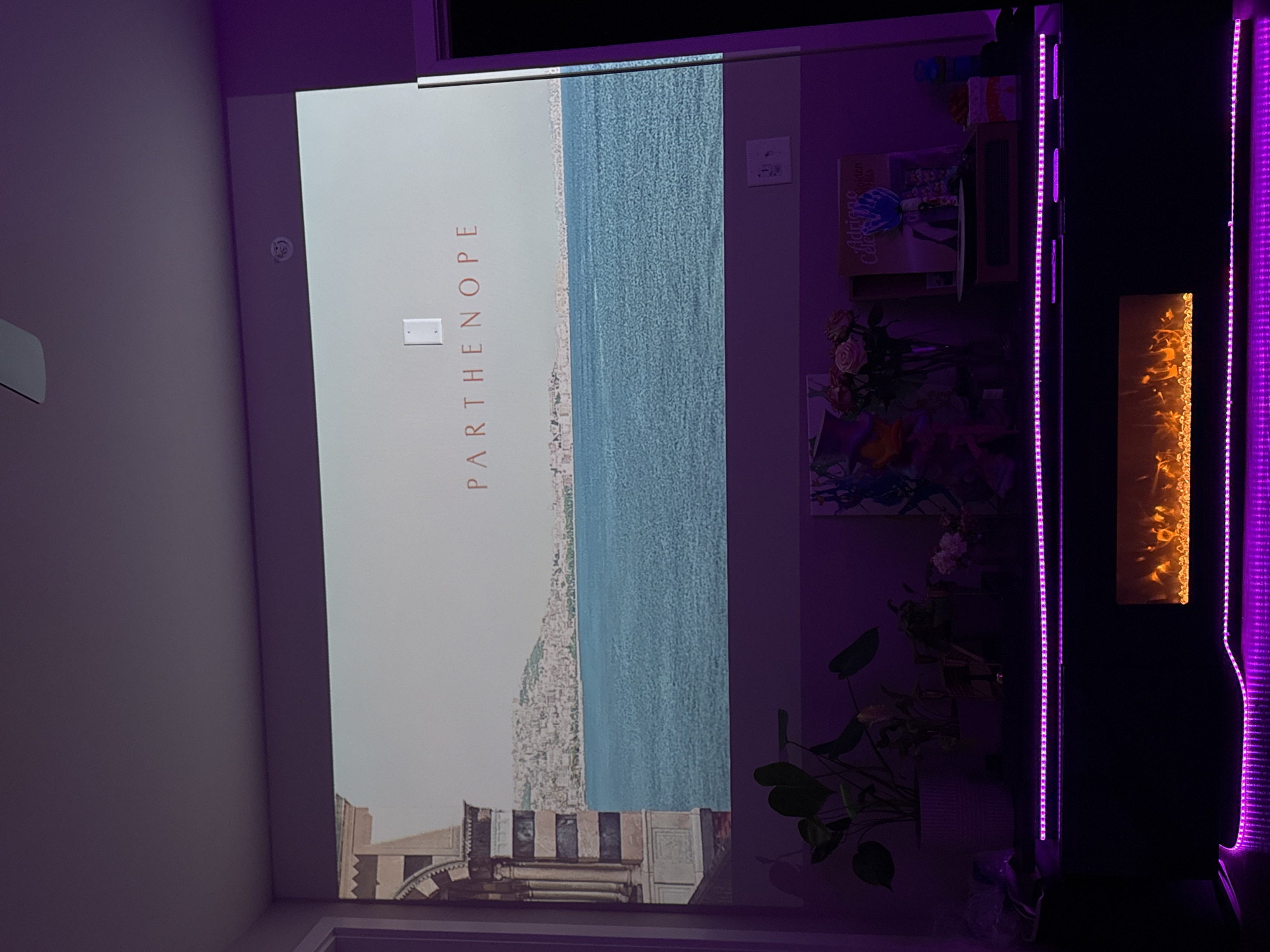 The image depicts a room interior with a focus on a large print of an artwork titled "Parthenope," showing a coastal city landscape. The print is positioned on a wall in what appears to be a residential setting. The city depicted in the artwork is situated along the coast, with a dense urban layout stretching along the shoreline and reaching into the hills. The artwork has a vertical orientation with the word Parthenope written vertically. The environment is illuminated by purple LED strip lights, giving a stylized appearance to the image. Other items in the room include plants, bookshelves and small ornamental decorations. The city in the background is located on the water and is not identifiable.