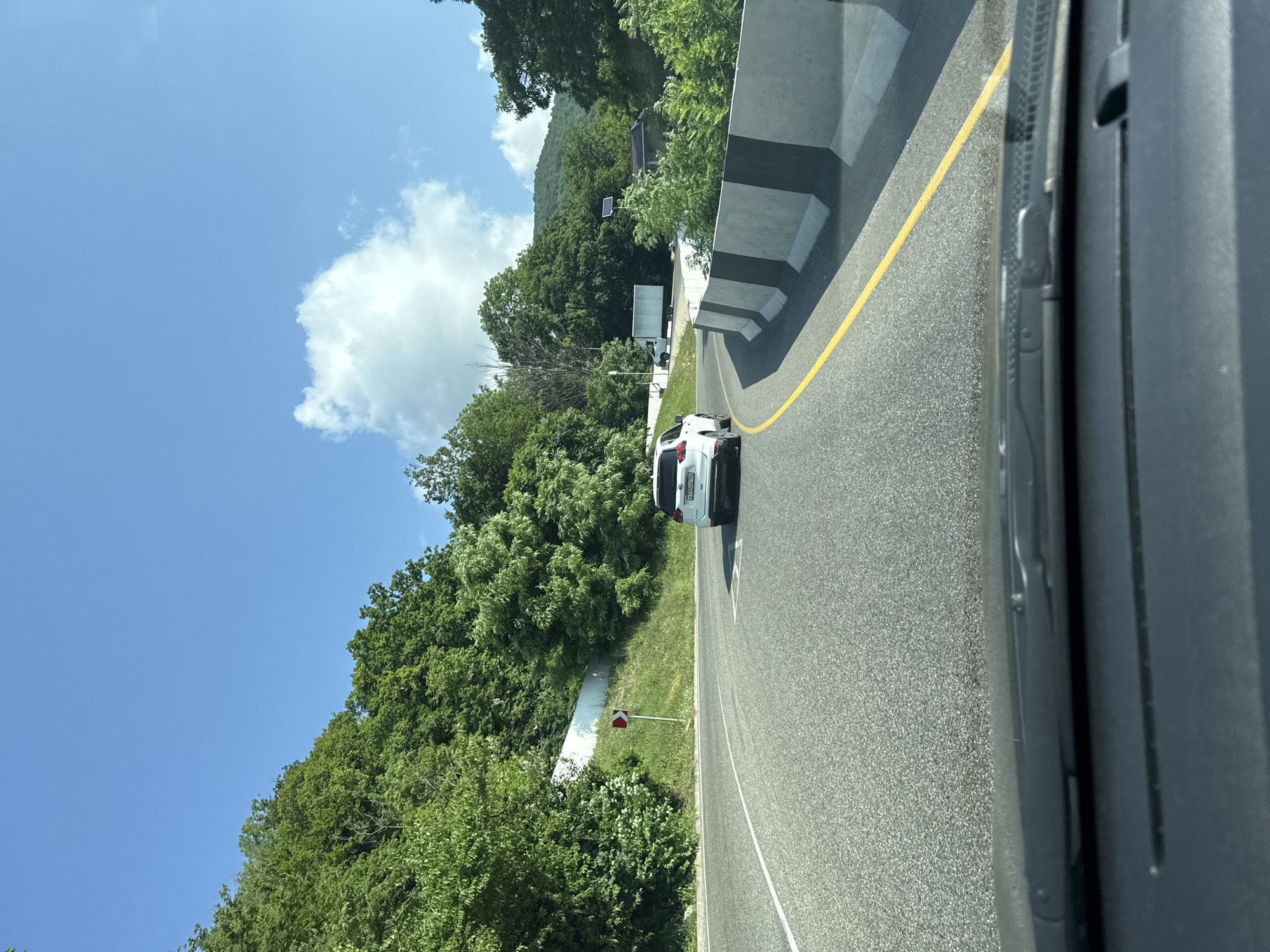 A white SUV is driving on a road with a yellow line. The road is surrounded by trees and hills, and there is a blue sky with white clouds. The image appears to be taken from inside another vehicle. The location is identified as Dederkoi, Russia.