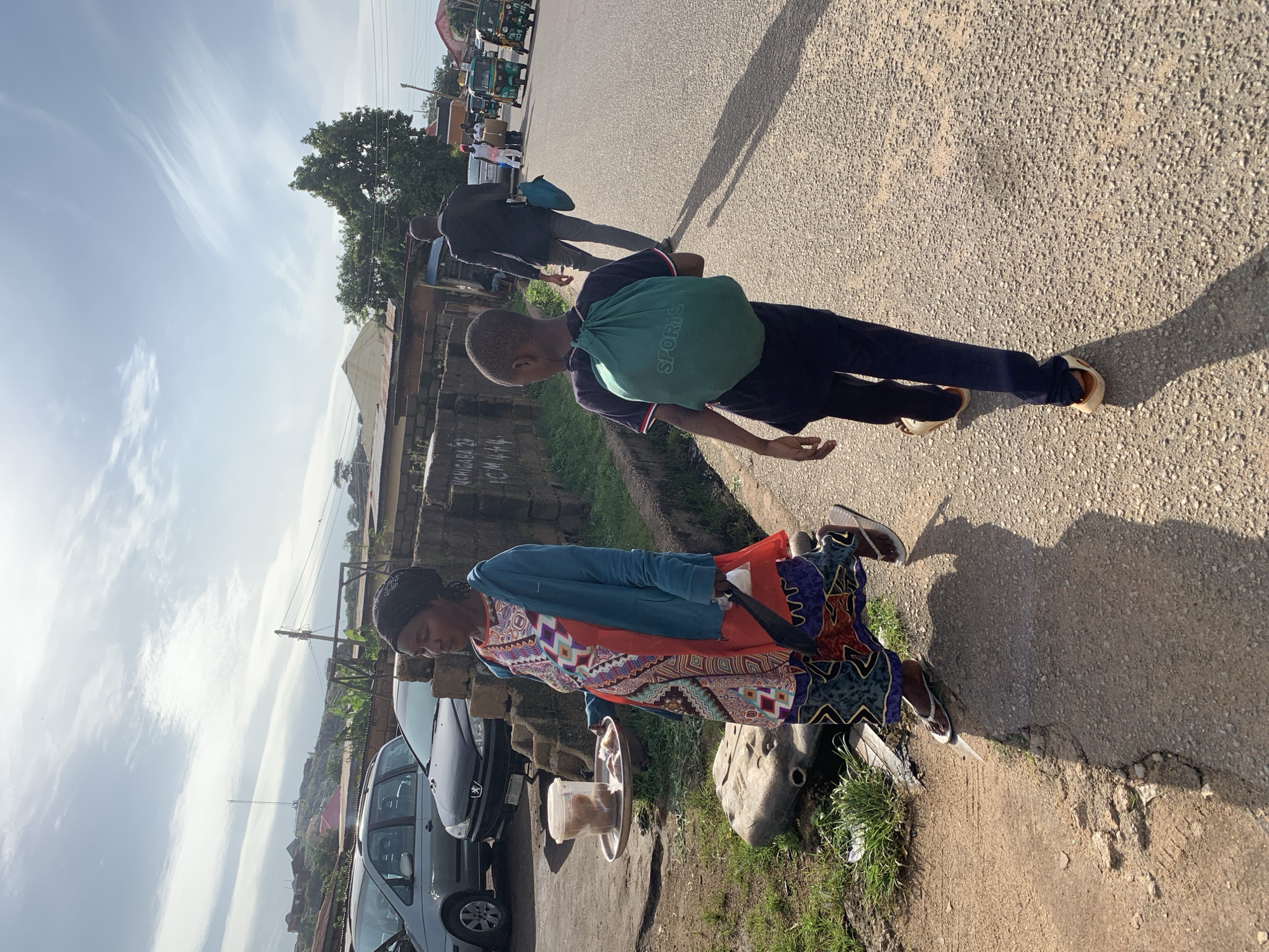 In Jos, Nigeria, an outdoor street scene unfolds under a partly cloudy sky. A woman walks along a paved road, carrying a tray of food in her left hand and a plastic bag in her right. She is dressed in a vibrant, patterned dress and a blue jacket, with flip-flops on her feet. A young boy walks ahead of her, carrying a green backpack with the word "SPORTS" printed on it. He wears dark pants and light-colored shoes. In the background, several vehicles, including cars and three-wheeled auto-rickshaws, are visible. The environment includes a dirt roadside with overgrown grass and a low wall constructed of bricks. The overall scene captures a moment of daily life on a street in Jos.