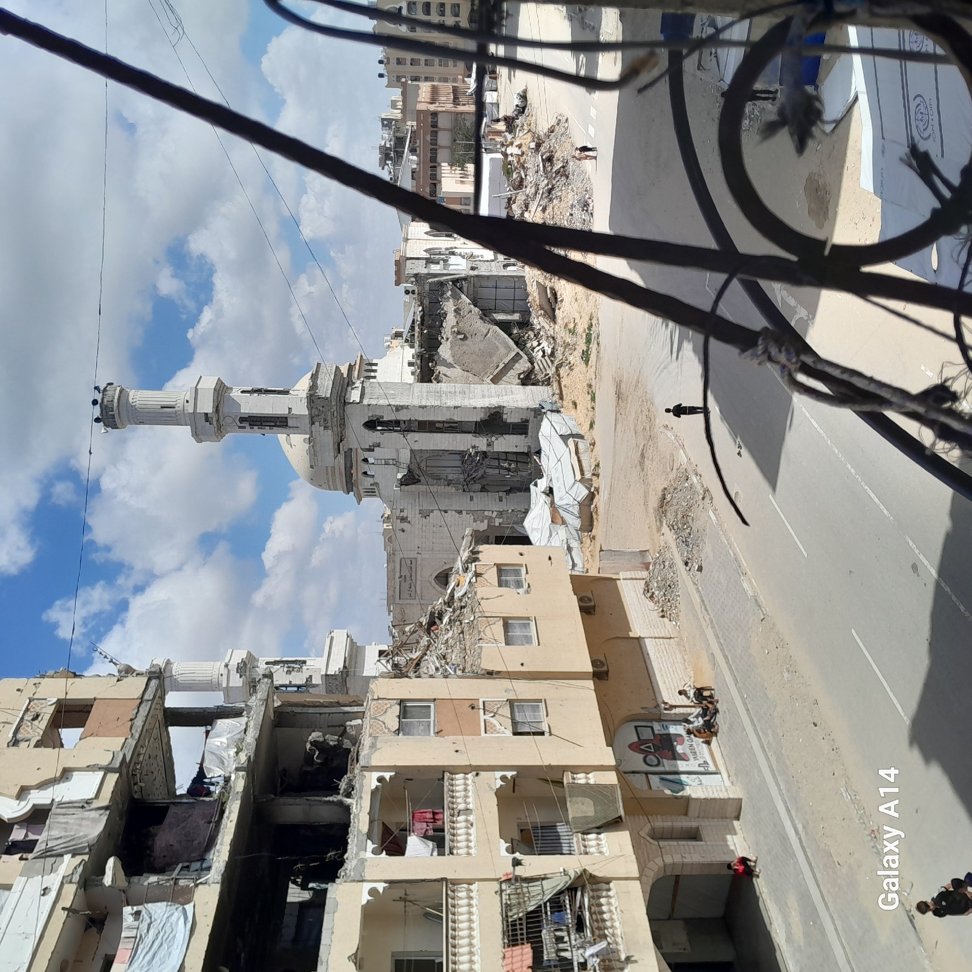 The image depicts a scene of urban devastation in Unknown, Unknown. A partially destroyed mosque with a minaret stands amidst damaged buildings. Debris is scattered along the street. A few individuals are visible; one person walks alone on the street. Several other individuals are gathered under the shadow of a destroyed building. The sky is blue with scattered clouds. Wires and a partial view of a structure obstruct the view.