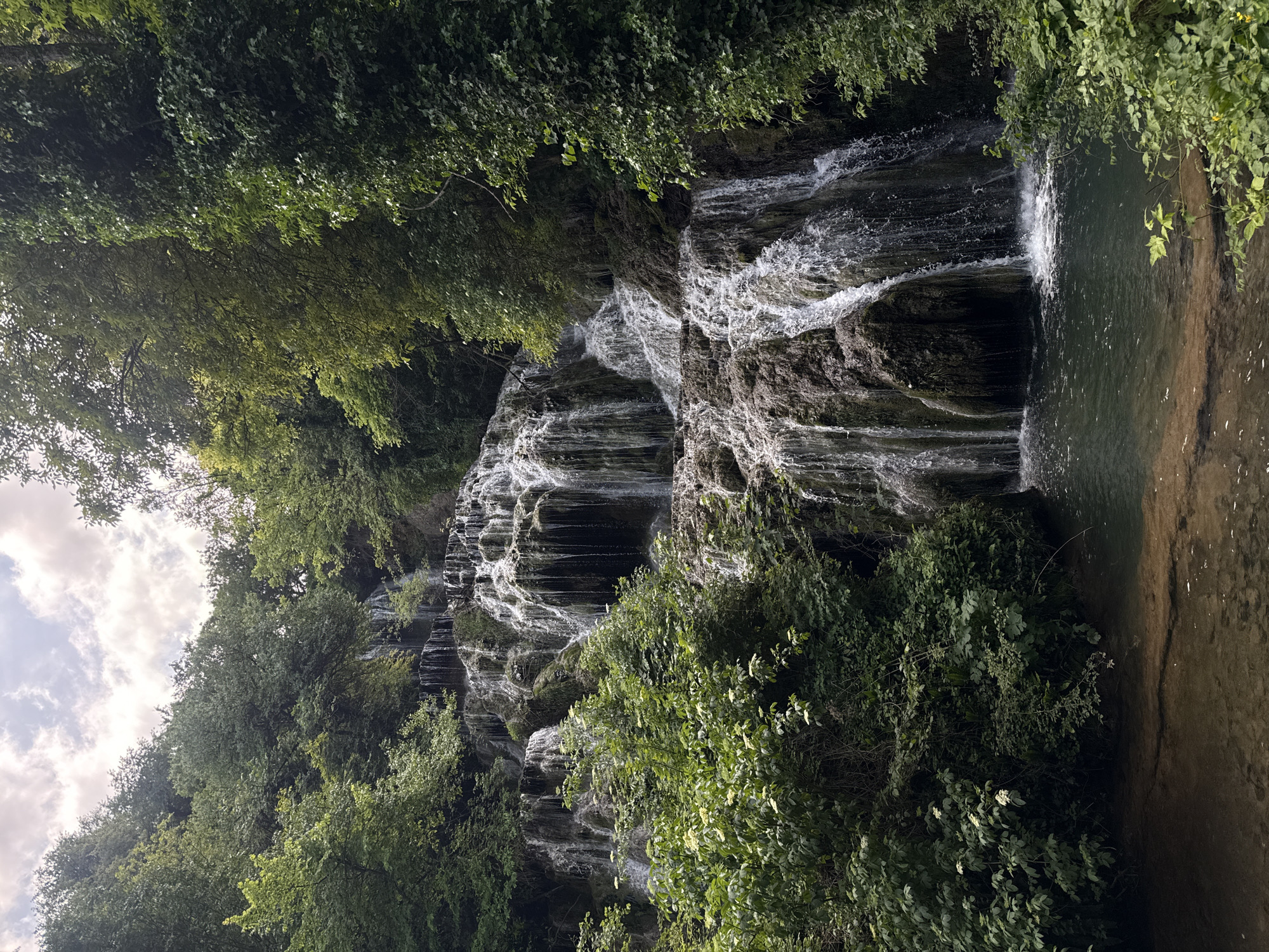 The image depicts a waterfall in Krushuna, Bulgaria. The water cascades down a series of rocky terraces, creating multiple small waterfalls. Lush green vegetation, including trees and bushes, flanks both sides of the waterfall. A pool of water is visible at the base of the falls on the right side of the image. Some sky with clouds can be seen peeking through the trees at the top left.