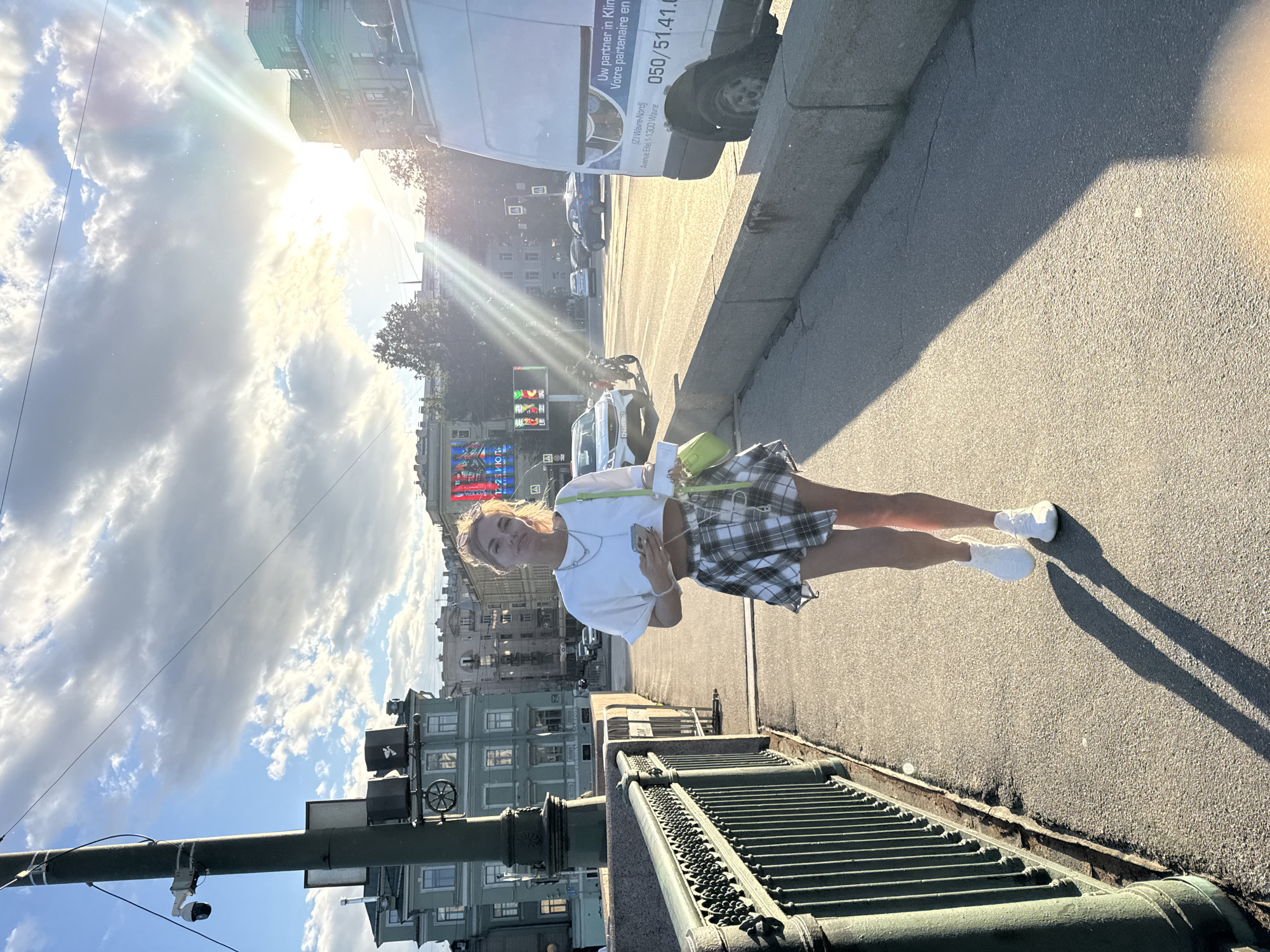 An athletic, blonde woman with a light tan walks toward the camera on a bridge sidewalk in Saint Petersburg, Russia, on a sunny day. She is wearing a white crop top, a plaid skirt, and white sneakers. She holds a mobile phone in her right hand and a bright green handbag in her left. The sidewalk is paved with asphalt, and a decorative green metal fence borders the left side of the bridge. In the background, buildings and trees line a street with moderate traffic, including cars and a motorcycle. The sun is setting, casting long shadows and creating lens flare. The sky is blue with scattered white clouds.