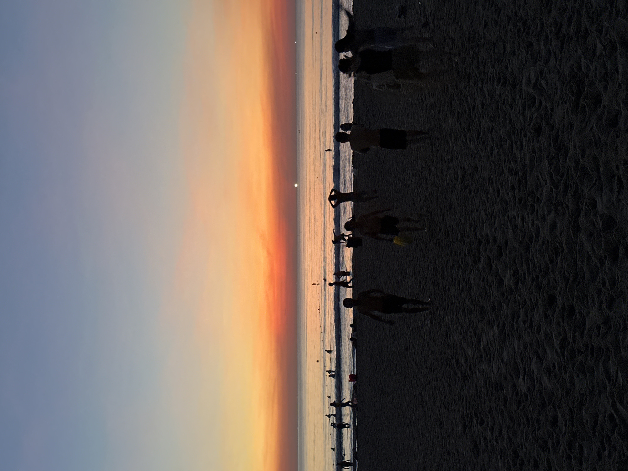 This media file shows a group of people on a sandy beach during what appears to be sunset or sunrise. The sky displays a gradient of colors, transitioning from a soft blue to warm oranges and yellows, suggesting it is either early morning or late evening. The sun itself is visible as a bright spot near the horizon. The sea is calm with gentle waves lapping at the shore.
The people are silhouetted against the bright sky and water, making it difficult to discern individual features. They appear to be casually walking or standing on the beach. Some are close to the water's edge, while others are further up on the sand. Their postures suggest a relaxed atmosphere. The sand has a textured appearance, with ripples indicating the presence of wind or receding water.
The scene is set at a beach, likely in Đà Nẵng, Vietnam, as per the location context. The overall mood is serene and tranquil, characteristic of a beach at dawn or dusk. There is no visible text in the image.