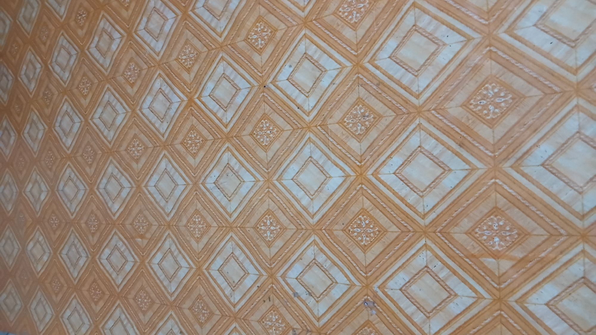 The image shows a close-up, high-angle shot of a floor with a diamond tile pattern. The tiles are predominantly light brown and off-white, with a repeating geometric design within each diamond. The pattern appears to be printed or embossed on a flat surface, possibly linoleum or vinyl flooring. There are some minor dark spots on the floor, which could be dirt or scuffs. The lighting suggests an indoor environment, with a slight glare visible on the surface, indicating it is a smooth material. The specific location in Prabumulih, Indonesia, is not discernible from the visual information provided in the image.