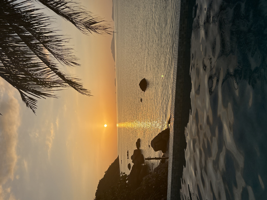 The scene depicts a sunset over the Andaman Sea from Ban Ko Li Pe, Thailand. In the immediate foreground, the rippling, dark blue water of a swimming pool is visible, bordered by a tiled edge consisting of small, square brown and blue tiles. Beyond the pool, the calm ocean extends, reflecting the golden light of the setting sun. A prominent, round, orange sun disc is low on the horizon, casting a bright, vertical glare across the water. To the left, a dark, rocky coastline with multiple large boulders rises from the water's edge. A slender, dark pole or post is vertically embedded near these rocks. Several isolated dark rocks are scattered within the ocean in the mid-distance. On the far left, a dark, vegetated landmass forms a silhouette against the sky. In the background, multiple smaller islands or landforms are visible on the distant horizon. The sky is a gradient of vibrant orange and yellow, transitioning to a lighter blue with faint clouds near the top. Silhouetted fronds of a palm tree frame the upper right portion of the image. No individuals are present.
