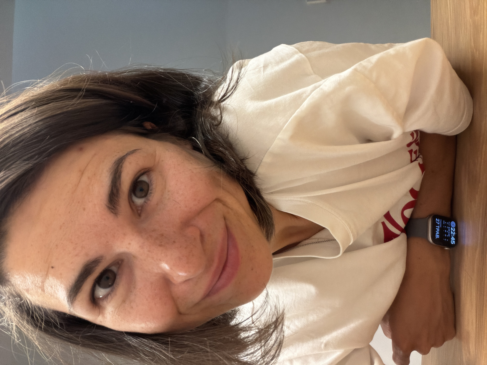 A woman with brown hair and freckles smiles at the camera. She is wearing a white t-shirt. Her smartwatch shows the time 22:45 and date 27 ТРАВ. She appears to be indoors. There is a light blue wall and a wooden surface in the background. Location in Budva, Montenegro cannot be determined from the given information.