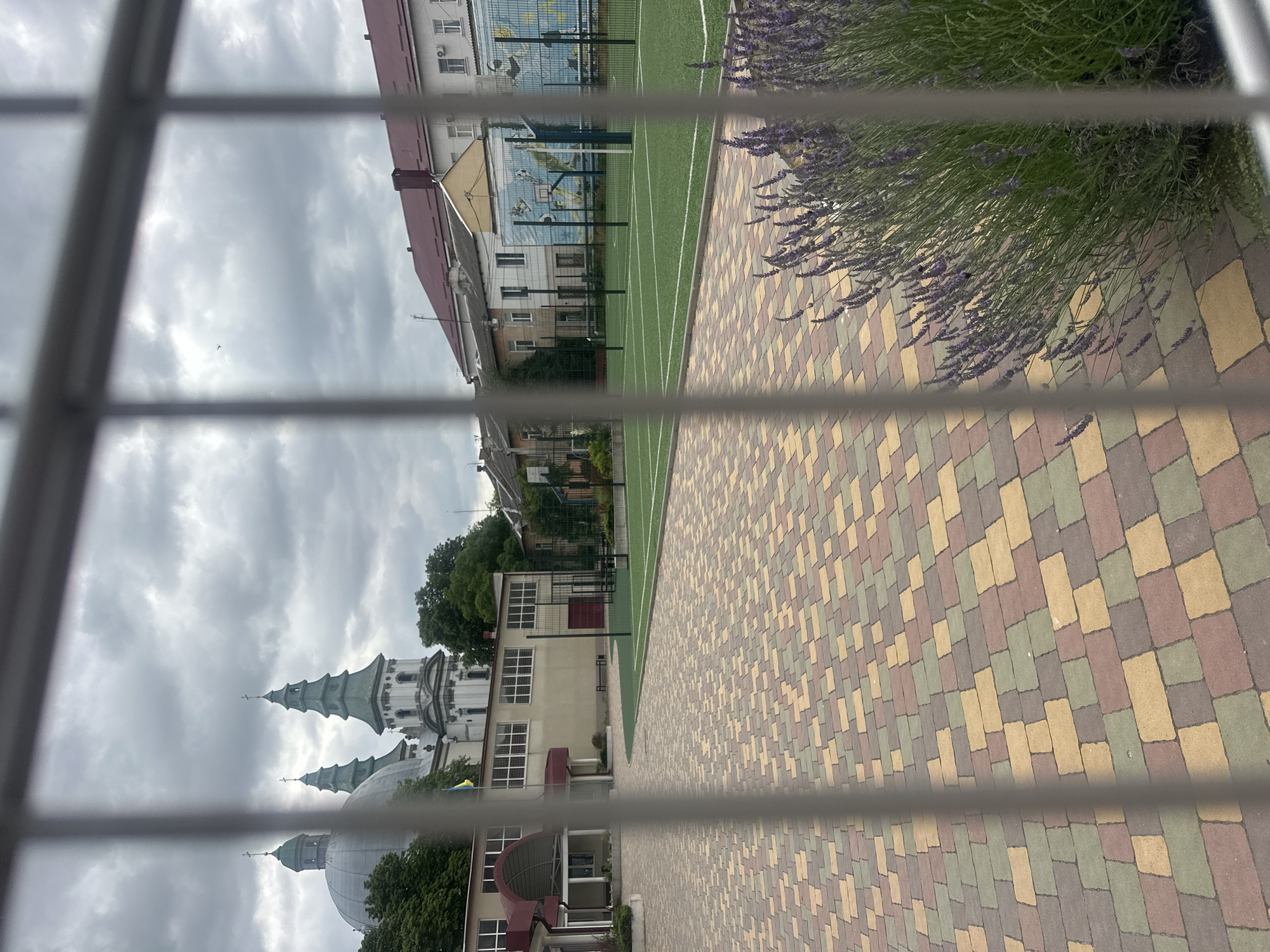The image shows an outdoor courtyard area in Ternopil, Ukraine, looking towards a sports field and adjacent buildings, with a prominent church visible in the background. The foreground is a paved area with multicolored interlocking bricks. To the right, a dense patch of lavender is in bloom. Beyond the paved area is a green artificial turf sports field with white markings, enclosed by a black chain-link fence. Two basketball hoops are visible on the far side of the field, mounted on colorful murals depicting sports scenes. In the mid-ground, several buildings are visible, including a light-colored school or institutional building with large windows and an arched entrance on the left. Further back, a Ukrainian flag is displayed on a flagpole. Dominating the background is a large, white church with multiple green domes and spires, characteristic of Orthodox Christian architecture. The sky is overcast with heavy, gray clouds. The scene is viewed through the bars of a light-colored metal fence, which partially obscures the view of the courtyard and sports field. No people are visible in the image.
