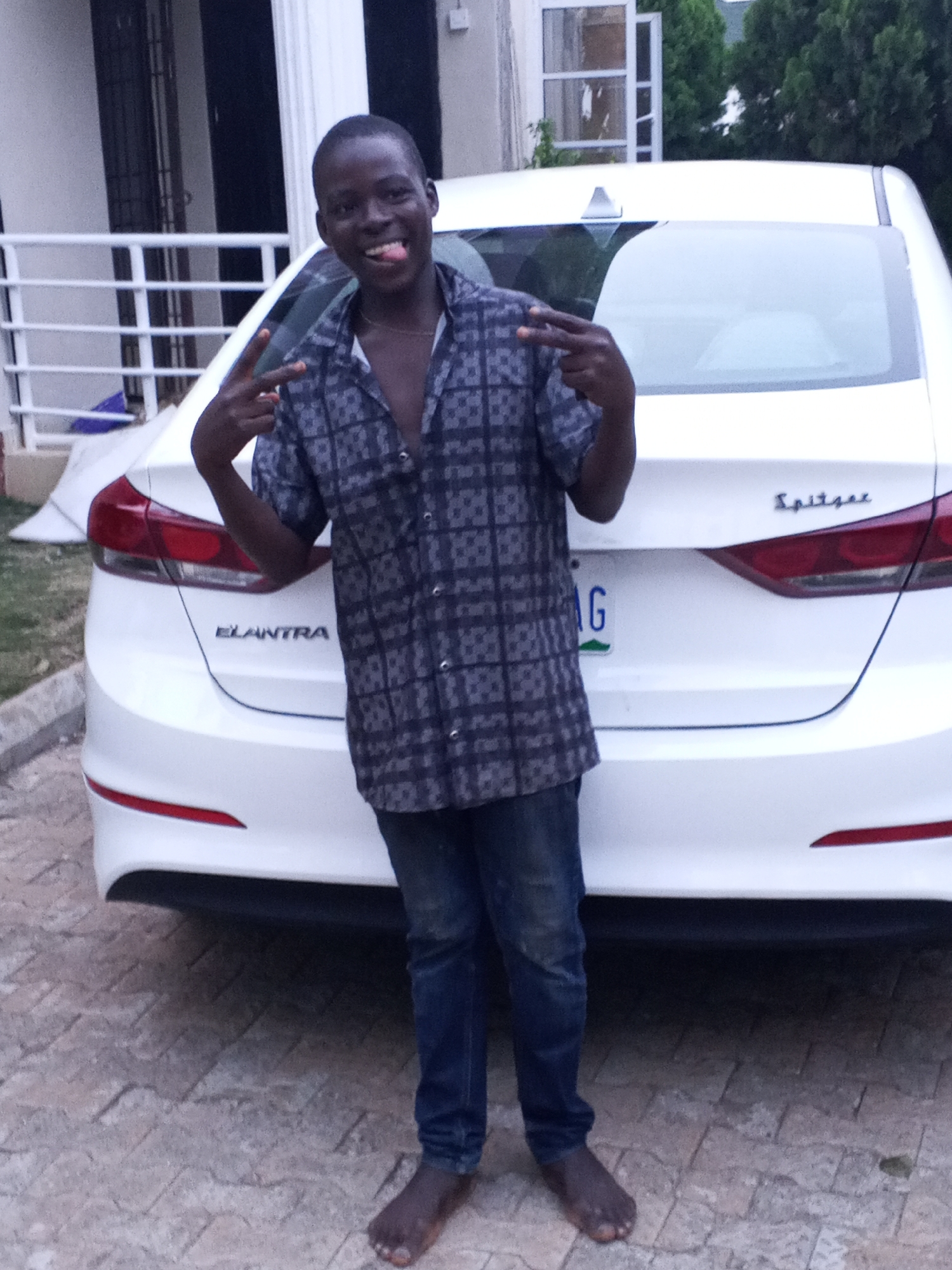 A young person is featured prominently in a full-length portrait, standing barefoot in front of a white Hyundai Elantra car, likely in a residential area of Jos, Nigeria. The individual, who appears to be male with dark skin and short hair, is smiling broadly with his tongue playfully sticking out and making peace signs with both hands. He is wearing a dark, patterned short-sleeved shirt, which is partially unbuttoned, and dark blue jeans.
The white Hyundai Elantra sedan is parked on paved ground composed of interlocking stones. The car's trunk clearly displays the "ELANTRA" model name on the left and "Spitze" (or a similar trim name) on the right side. A partial license plate, featuring the letter "G" against a green and white background, is visible below the "Spitze" text.
In the background, a building with white railings and dark window frames can be seen, suggesting a house or similar structure. Green foliage from trees or bushes is visible behind the car on the right. The lighting indicates it is daytime, possibly late afternoon or early evening, creating a generally bright but diffused illumination. The overall impression is one of a cheerful and lighthearted moment.