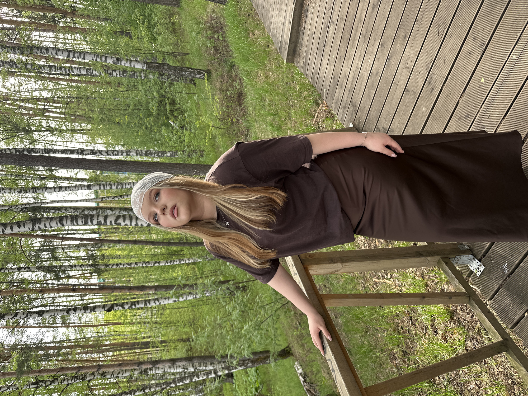 A young woman is standing on a wooden deck with a wooded area as the backdrop, seemingly in Chaikovsky, Russia. The woman is wearing a brown dress and a light-colored headband. She is leaning on a wooden railing, and her gaze is directed towards the viewer. The background consists of birch trees with green foliage, typical of a forest environment. The wooden deck has parallel planks running horizontally. Sunlight is filtered through the trees, creating shadows and highlights within the scene.