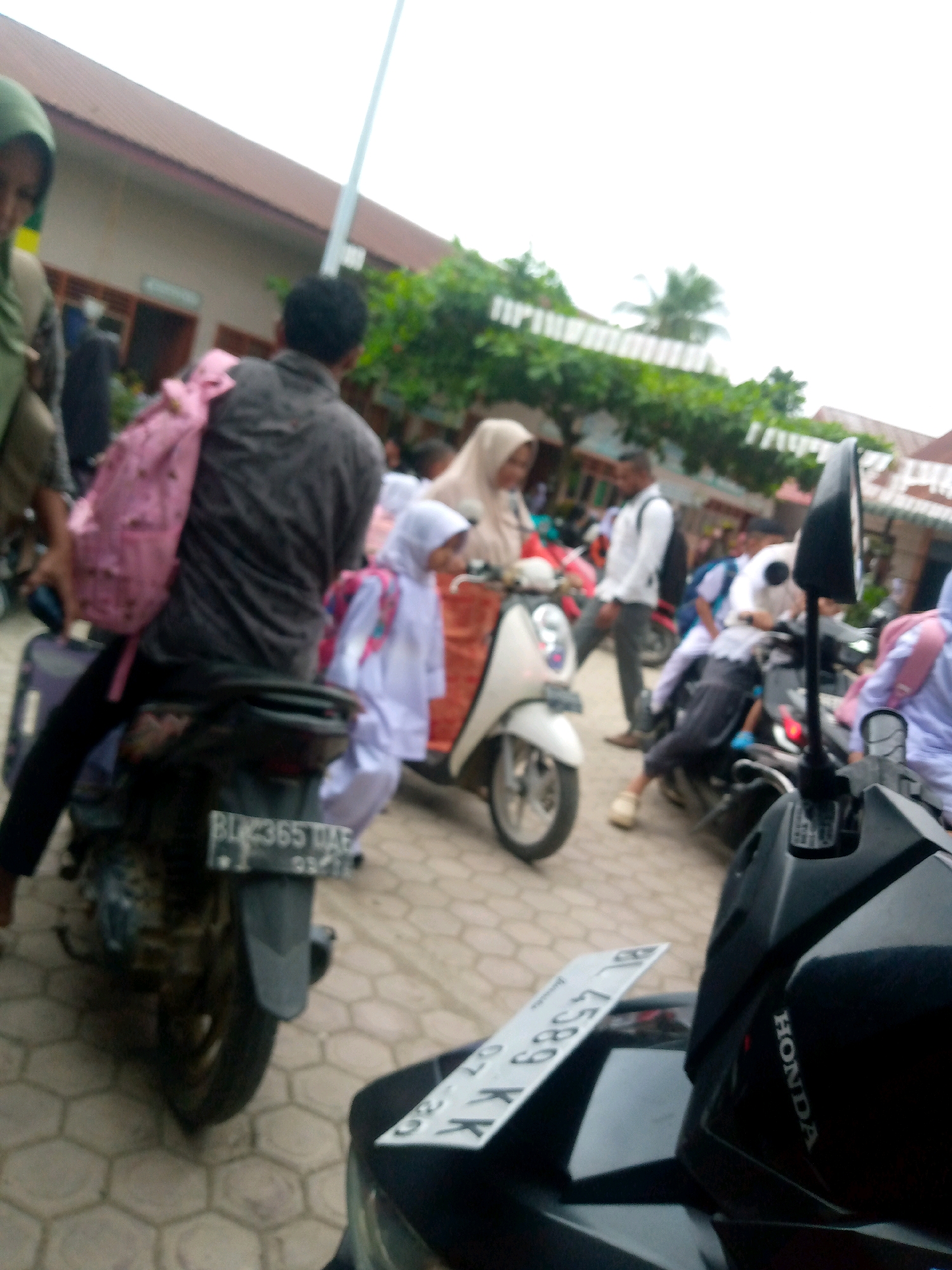 A low-angle, slightly blurry, outdoor, daylight, wide shot captures a busy scene with several people on and around motorcycles. The ground is paved with hexagonal tiles. In the foreground, the rear of a black motorcycle is visible, with a license plate reading "BU 4589 KX". To the left of this, a person in a dark grey shirt and black pants sits on another motorcycle, a large pink backpack with a floral pattern rests on their back, and a dark suitcase is attached to the side. To the right of the foreground motorcycle, a black mirror with a red accent reflects the scene.
In the mid-ground, a person in a white headscarf and beige robe is seated on a white motorcycle. To their right, a person in a white shirt and grey pants walks away from the camera, carrying a white bag. Further back, a person in a white uniform with a blue backpack is seated on a motorcycle. Another person in a white headscarf and light purple top is also on a motorcycle. To the far left, a person wearing a green headscarf is partially visible. The background shows a building with a reddish-brown roof and some trees. The overall impression is a bustling transportation hub, possibly near a school given the attire of some individuals. The location is identified as Mesjid, Indonesia.