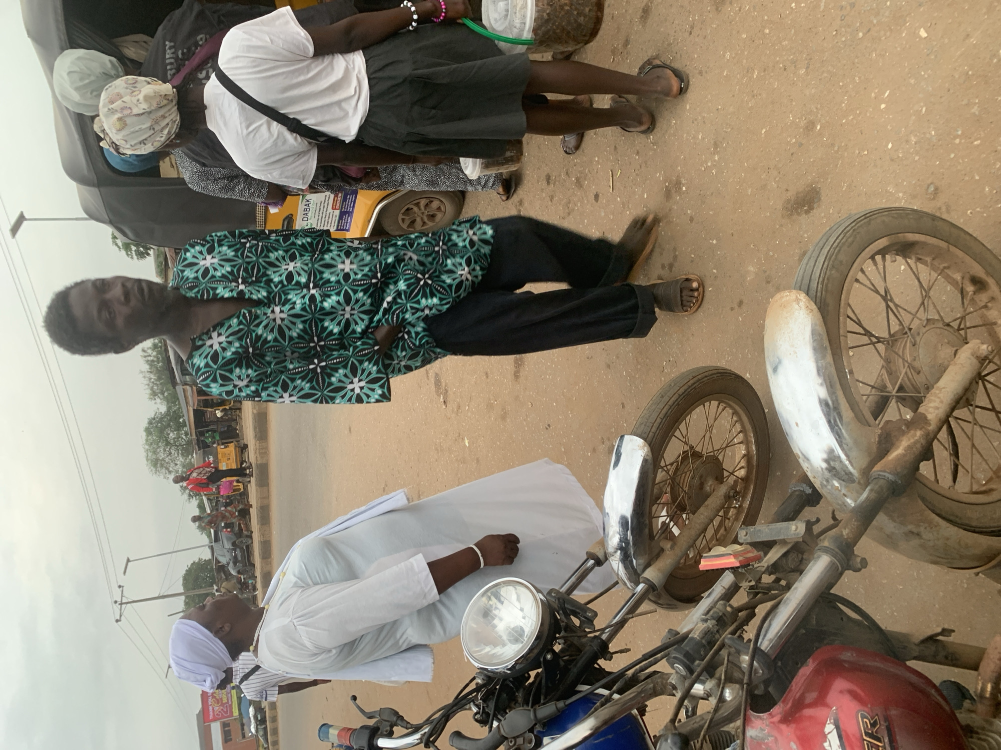 In the city of Shagamu, Nigeria, a group of people are gathered near a yellow motorized tricycle, likely a "keke napep." In the foreground, two motorcycles are parked, one red and one blue, showing signs of wear and tear. A man in a patterned green and black shirt and dark pants stands looking ahead, his feet bare in sandals. To his right, a woman in a white robe and headscarf, possibly a member of a religious group, stands with her arm bent. Further back, another woman in a white t-shirt and black skirt is holding two buckets, one green and one clear plastic, filled with what appears to be produce or goods. Behind her, other individuals are visible in and around the keke napep. The ground is a dusty, reddish-brown dirt, and the sky is overcast. The scene suggests a busy roadside or market area.