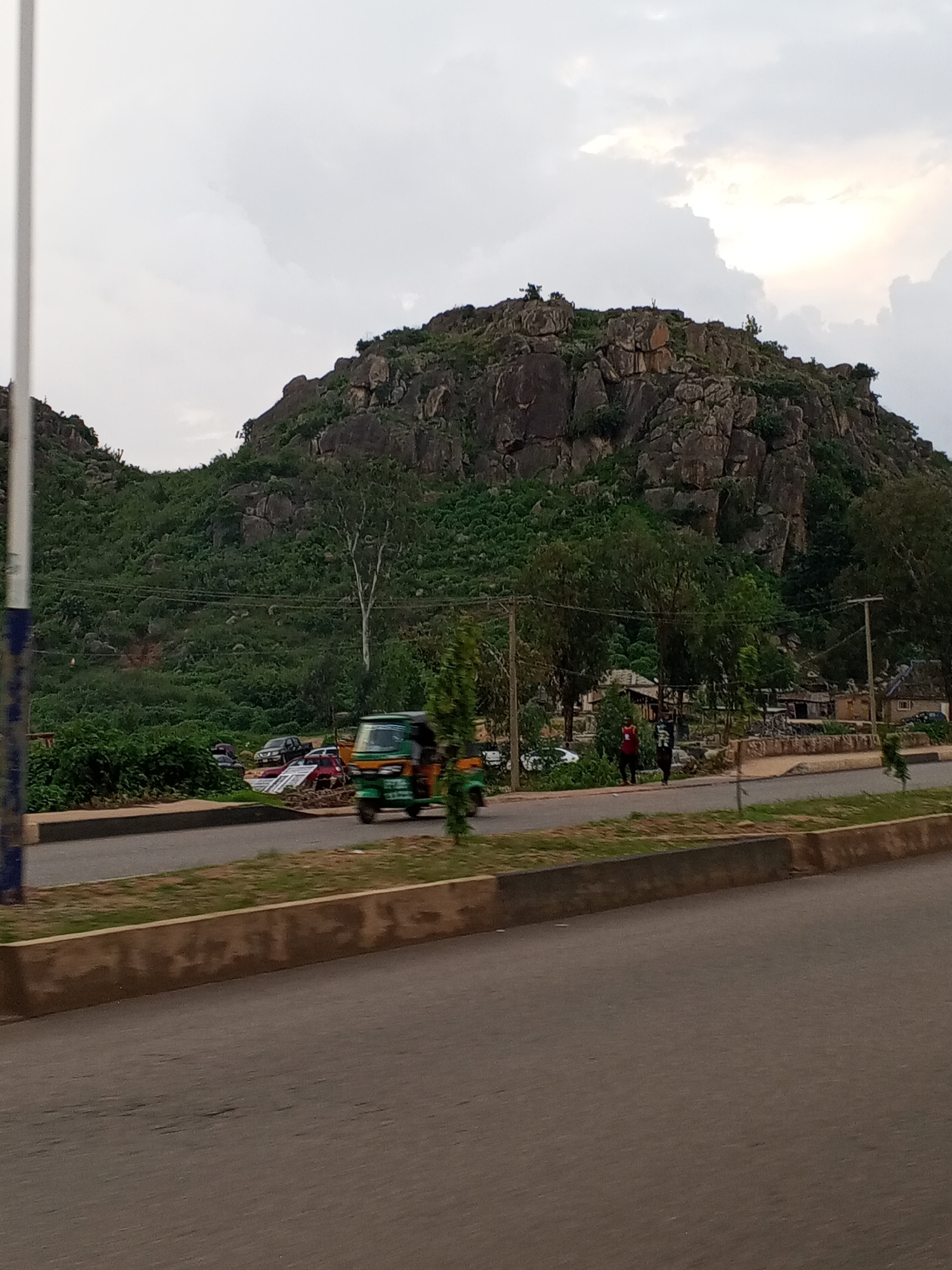 In Gura Top, Nigeria, a road runs parallel to a densely vegetated mountainside. A weathered, light-brown concrete barrier with a darker top edge separates the road from a strip of grass and dirt. Beyond the barrier, several vehicles are parked, including a green auto-rickshaw in motion, a red sedan, and two dark-colored cars. The auto-rickshaw is in the foreground, moving from left to right. The mountainside behind the vehicles is covered in green foliage and interspersed with large, dark grey rocks. A tall, slender tree with a white trunk stands near the center of the mountainside. The sky above is overcast with large, diffused clouds. Two figures are visible standing on a small dirt area near the parked cars.