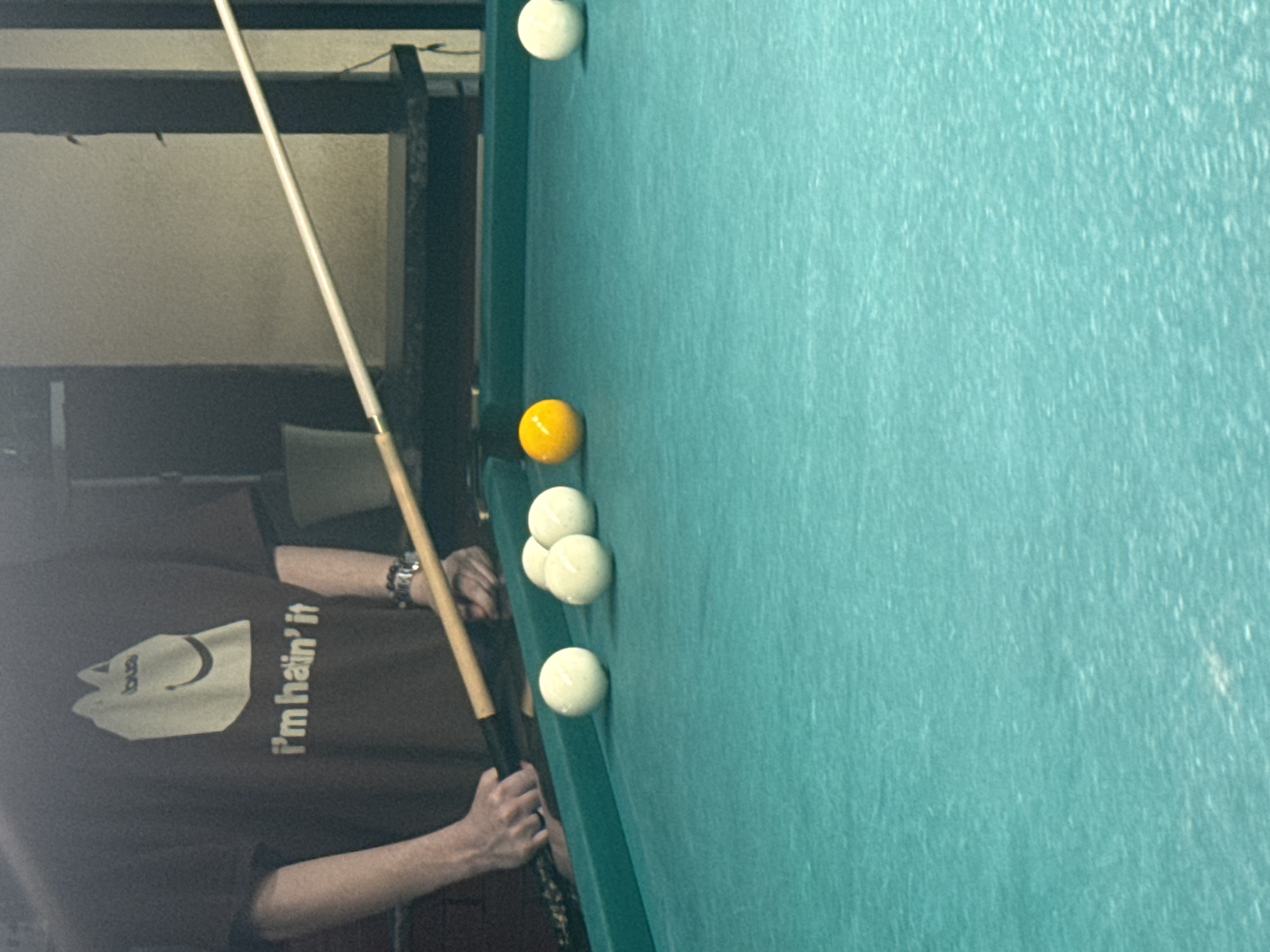 A person in a brown shirt with the text "I'm hatin' it" is playing billiards on a green felt table. There are six balls on the table: one yellow, one at the edge of the table, and four clustered close together. The person is holding a pool cue and appears to be lining up a shot. The location is likely indoors, but no specific location information for Moscow, Russia is present.