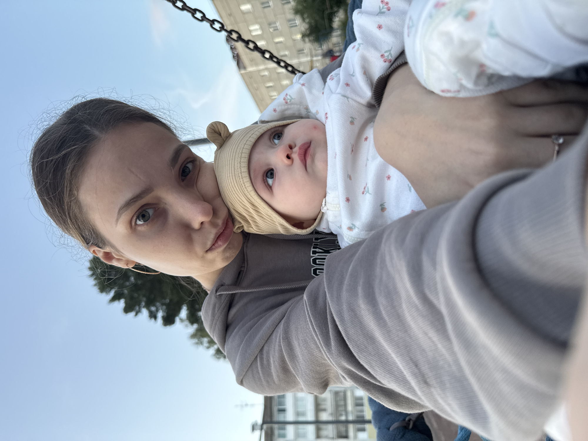 In Noviy, Russia, an adult female with light brown hair, wearing a grey hooded sweatshirt, is holding an infant. The infant is wearing a light beige hat with bear ears and a white onesie with a floral pattern. They appear to be on a swing, as indicated by the visible chains. The background shows a residential building and trees under a clear sky.