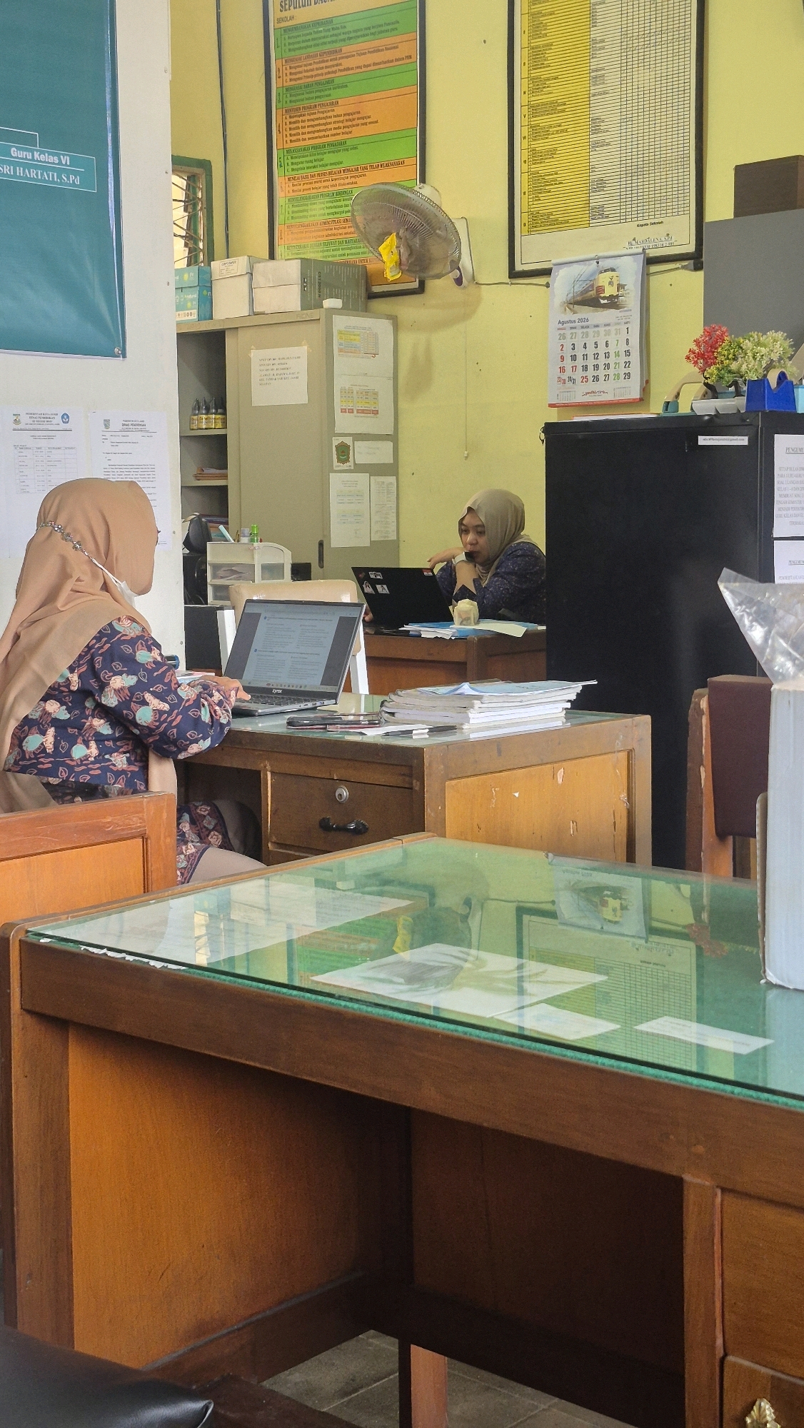 Two women are seated at desks in what appears to be an office or classroom environment. The woman on the left is wearing a light brown hijab and a blue, patterned top. She is looking at a laptop placed on her desk. The woman on the right, also wearing a hijab, is wearing a dark, patterned top and is looking towards her laptop. Papers and books are visible on both desks. In the background, there are filing cabinets, posters on the wall, and a calendar for August 2026. A fan is mounted on the wall. The overall scene suggests a workspace where administrative or educational tasks are being performed.