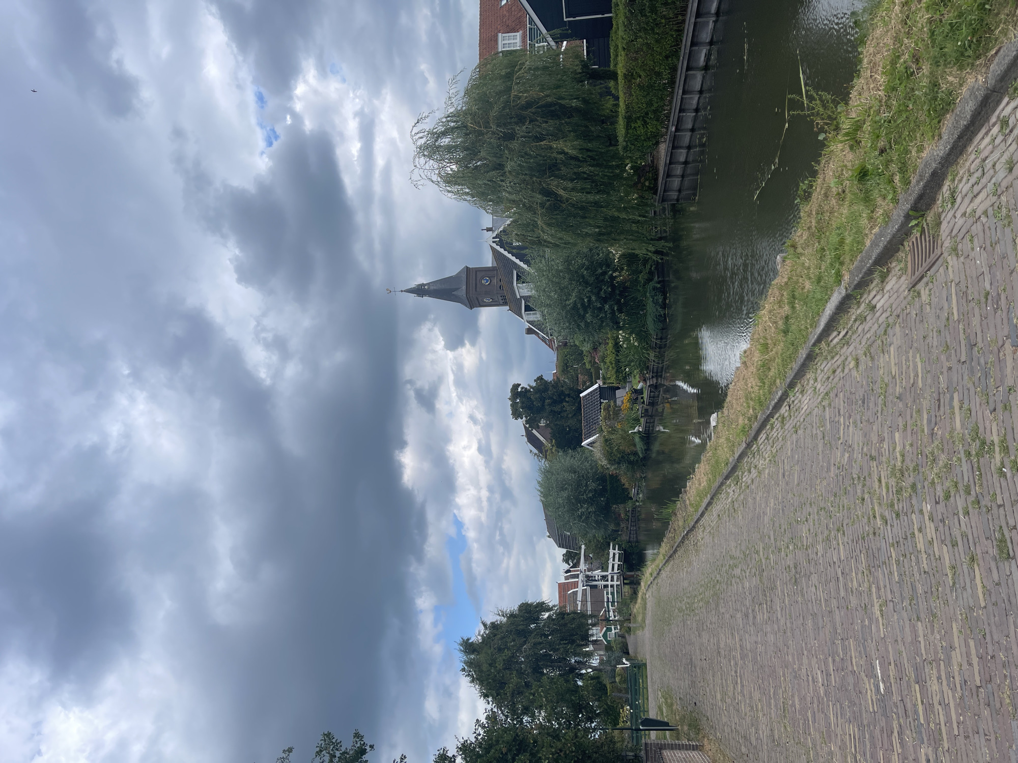 The image depicts a canal-side pathway in the city of Marken, Netherlands. The path is paved with red bricks and lined with grass. On the right side of the path is a canal with dark water reflecting the cloudy sky. Along the canal are houses with dark siding and red roofs, some of which have white trim. A church with a tall, dark steeple and a clock is visible in the background. Several trees and lush green vegetation are present, including a weeping willow tree on the right. The sky is overcast with heavy gray clouds, interspersed with some lighter patches. The overall impression is of a peaceful, somewhat moody, waterside scene in a Dutch village.