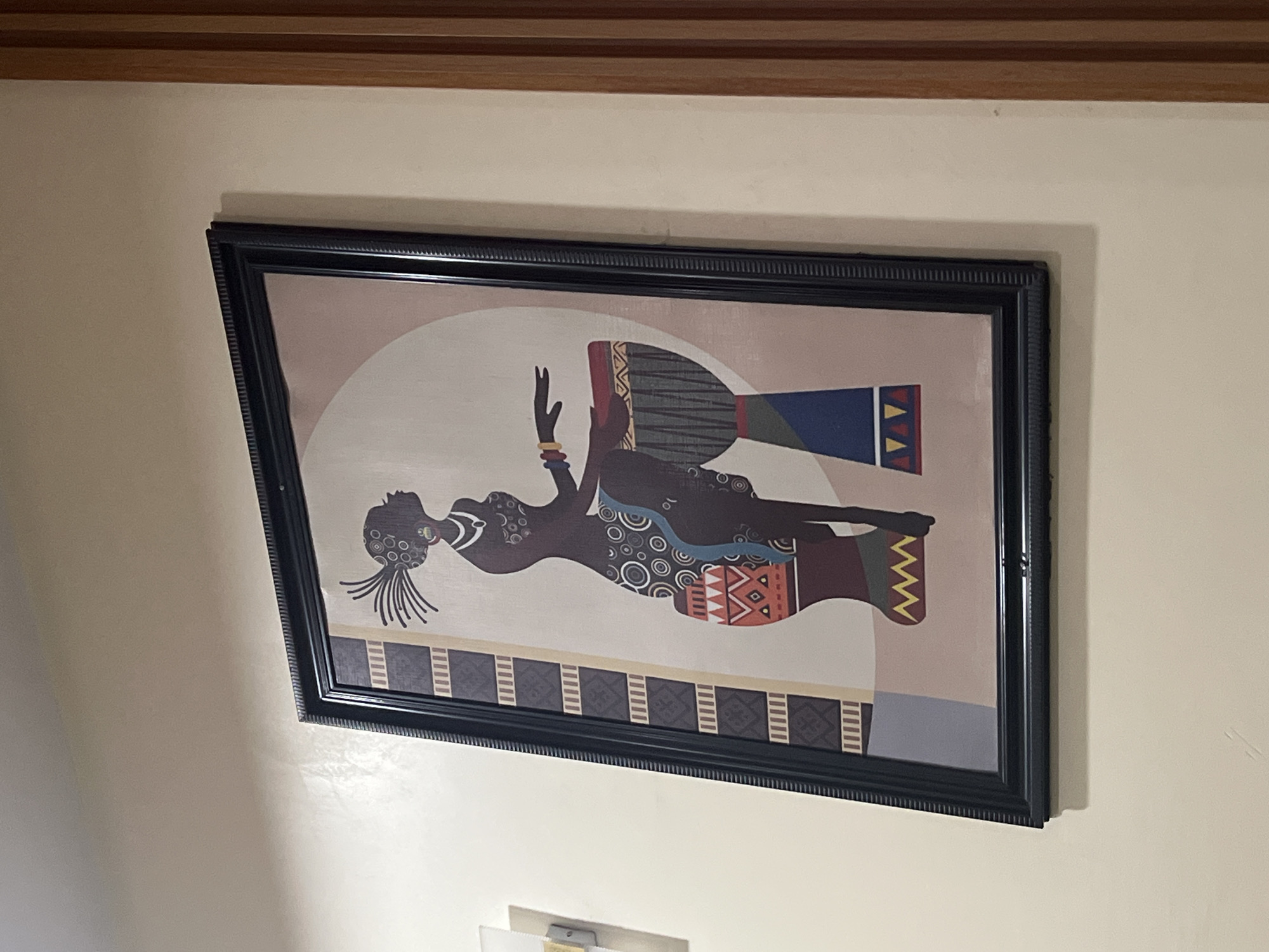 The image features a framed, stylized painting hanging on a plain, light beige wall. The original image is rotated 90 degrees counter-clockwise; in its correct upright orientation, the painting depicts an African-inspired scene.
The central subject of the artwork is a slender, stylized female figure, rendered predominantly as a dark silhouette. She exhibits intricate circular patterns on her torso and a distinctive, radiating hairstyle or headdress. Her arms are raised, and one hand, shown in a lighter skin tone, is adorned with multiple gold-colored bangles. She appears to be in a reclined or seated pose, looking towards the left. Positioned behind and slightly above her is a large, colorful drum, possibly a djembe, featuring bold geometric patterns in hues of red, blue, yellow, and dark grey. The painting's background is a soft beige, with a lighter, almost white, circular area behind the figure and drum. A decorative border with abstract patterns runs along the bottom edge of the canvas.
The painting is enclosed in a thick, black frame, which boasts a textured, possibly carved, pattern along its inner and outer borders. Above the painting, a wooden architectural trim, composed of two parallel strips, marks the junction between the wall and an unseen ceiling or another wall. A small, indistinct white object, possibly a light fixture or another wall-mounted item, is partially visible on the wall below the painting.
The scene is static, depicting no specific activity or event beyond the posed subject within the artwork. The setting is indoors. The stylized African aesthetic of the painting is culturally relevant, consistent with the provided location context of Gwarinpa, Nigeria. There is no visible text in the image.
