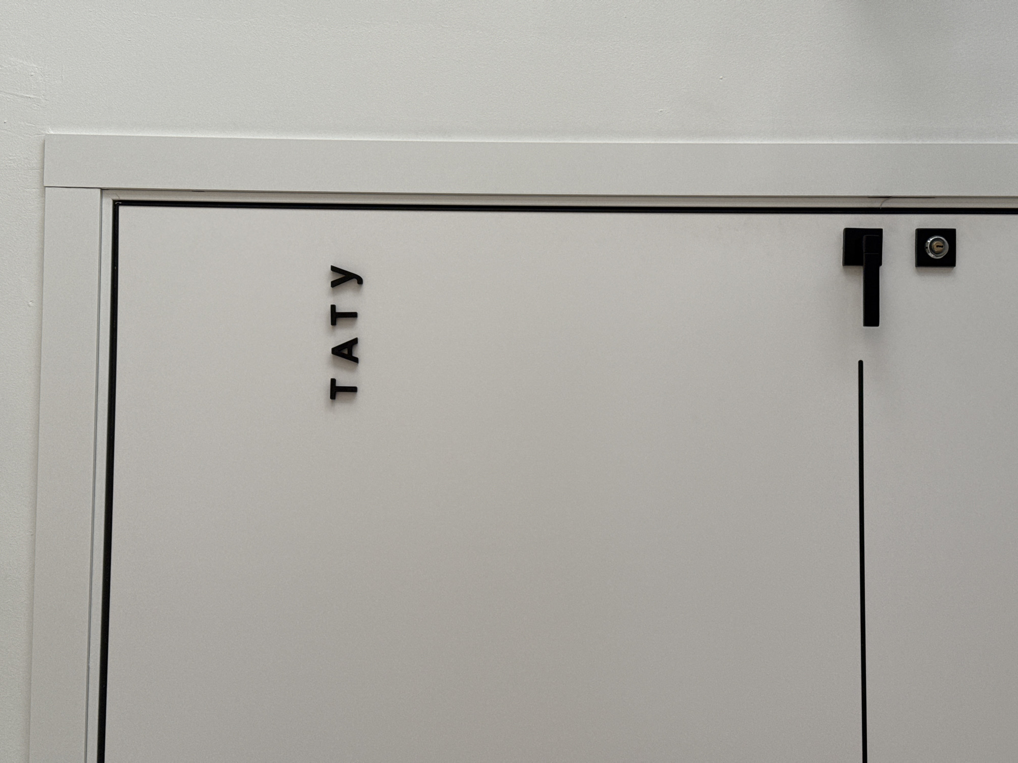 The image shows a white door with black lettering "TATY" vertically aligned on the left side. A black handle and a small round lock are on the right side of the door, along with a black vertical line running from the bottom of the handle to the bottom edge of the door. The door is framed by a white border. The location of the image is not given, but it has lettering that could represent the Russian pop duo t.A.T.u., with the city Krasnoyarsk as an optional location. No people are visible.