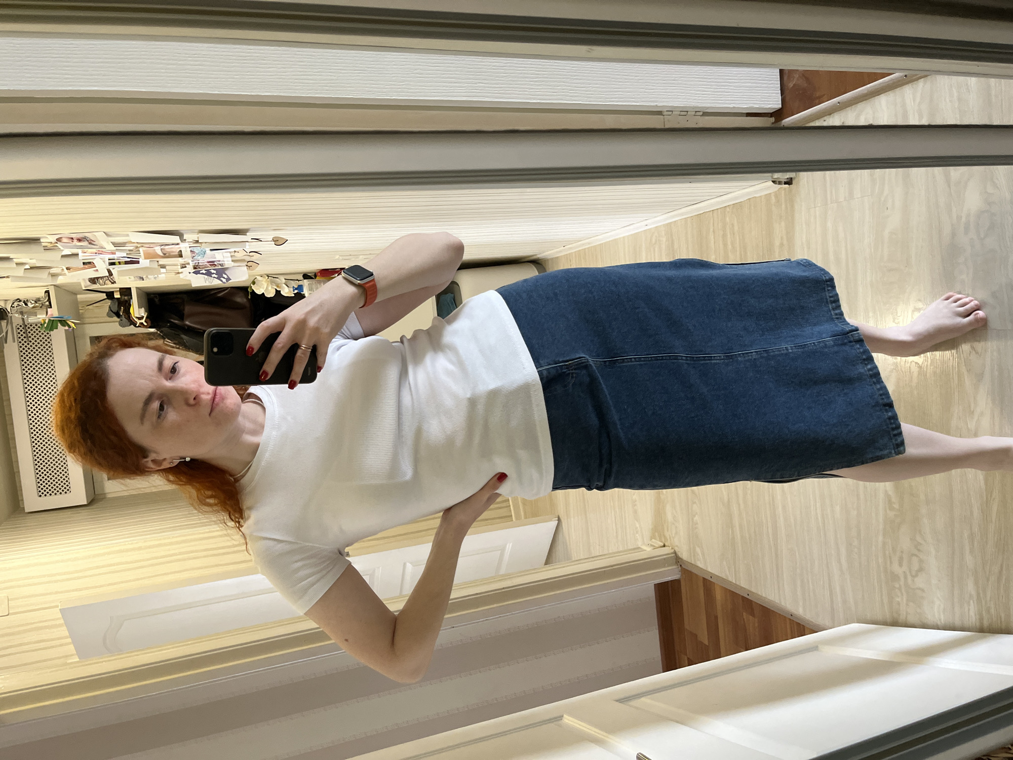 A woman is standing in what appears to be a room with light wood-patterned flooring. She is taking a photo of herself in a mirror. She is wearing a white t-shirt and a blue denim skirt. She is barefoot. The mirror is framed with a multi-layered white molding. Behind her in the reflection are a variety of items attached to a wall, including photographs and possibly decorative items. A white radiator cover is visible in the reflection. There is a white door frame in the lower portion of the reflection. The location and its connection to Chaikovsky, Russia, cannot be determined from the image.