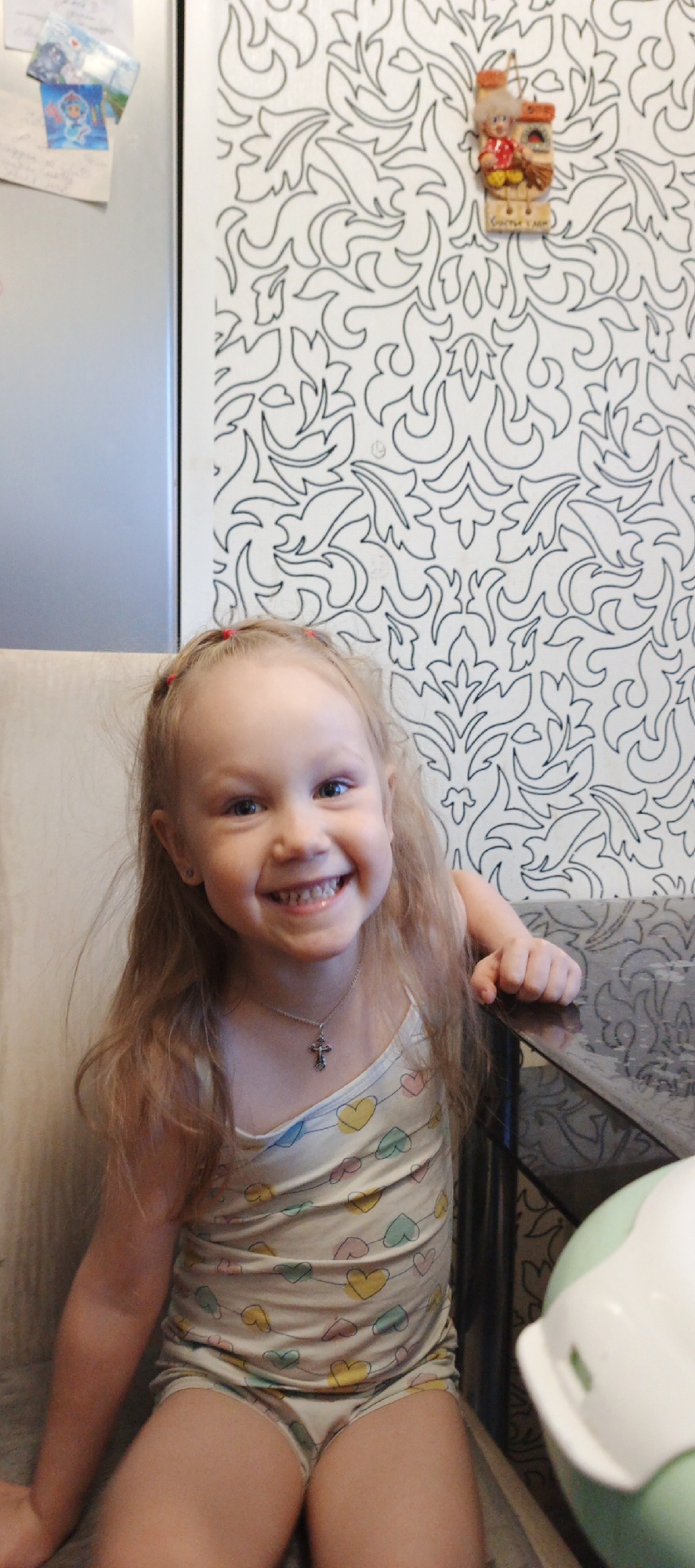 A young child, approximately 3-5 years old, with light brown hair and blue eyes, is seated indoors, smiling directly at the camera. The setting appears to be a residential kitchen or dining area in Chaikovsky, Russia. The child wears a light-colored tank top and matching bottoms featuring multicolored heart patterns, a silver-colored chain with a crucifix pendant, and small stud earrings. Her left hand rests on a dark, reflective table surface, which displays a subtle etched pattern. To the left, a portion of a light-colored upholstered chair or bench is visible. The background consists of a metallic grey refrigerator on the upper left, adorned with several attached items, including a picture resembling Elsa from Disney's Frozen and a handwritten note. The main wall surface is white with a repeating pattern of dark, stylized floral or scroll-like designs. A small decorative wall hanging, depicting a figure in traditional Russian attire next to a house-like structure, with the Cyrillic inscription "СЧАСТЬЯ В ДОМ" (Happiness to the home), is mounted on this patterned wall. A light green and white object, resembling a child's potty, is partially visible in the bottom right quadrant of the image.