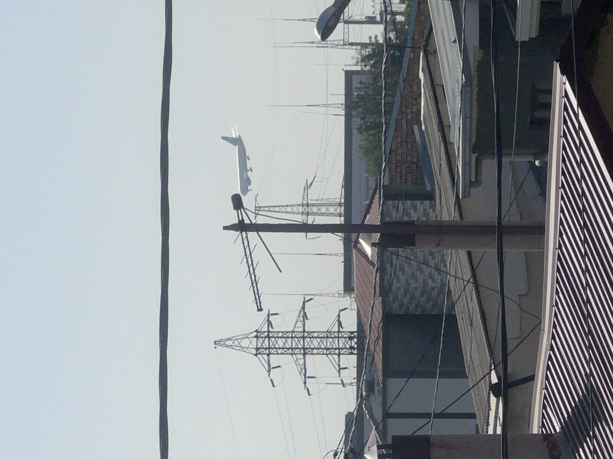A passenger airplane is captured in flight against a hazy, light blue sky in an urban environment. The airplane is white and appears to be a Boeing 737 or similar narrow-body aircraft, with its landing gear deployed, suggesting it is either approaching for landing or has just taken off. Below the aircraft, a cluster of power line towers, characterized by their lattice metal structures and numerous wires, dominates the mid-ground. These towers are interspersed with various low-rise buildings, some with visible corrugated metal roofs, and brickwork. In the foreground, more power lines crisscross the frame, obscuring parts of the scene. The overall impression is one of an industrial or residential area situated near an airport.
