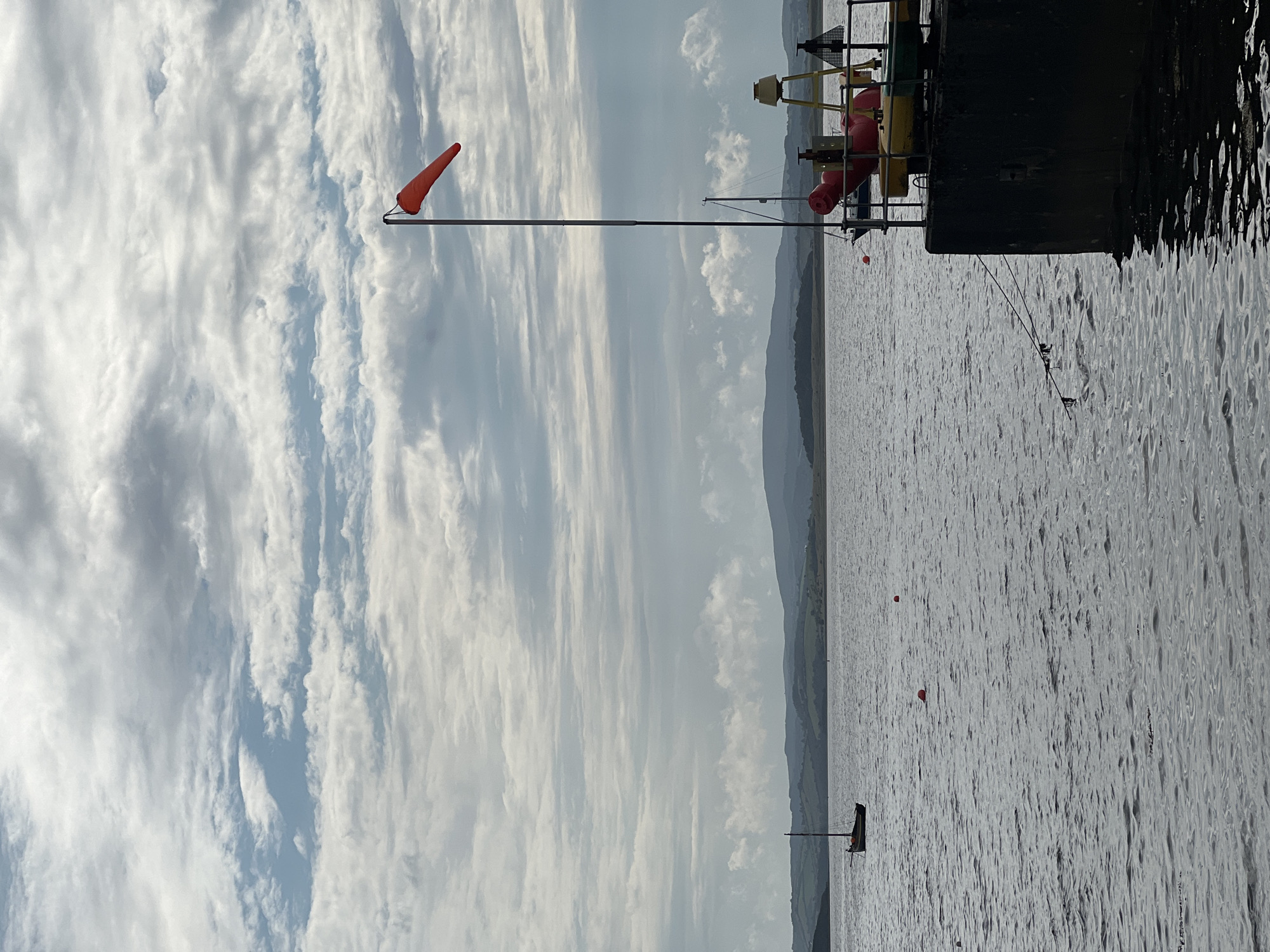 The image depicts a body of water with a pier in the foreground. A single sailboat is visible in the distance on the left. A tall pole with a red windsock is erected on the pier. Several buoys are scattered across the water. The pier itself is concrete and has railings. The background shows a distant, hilly coastline under a cloudy sky. The scene suggests a maritime or harbor environment.