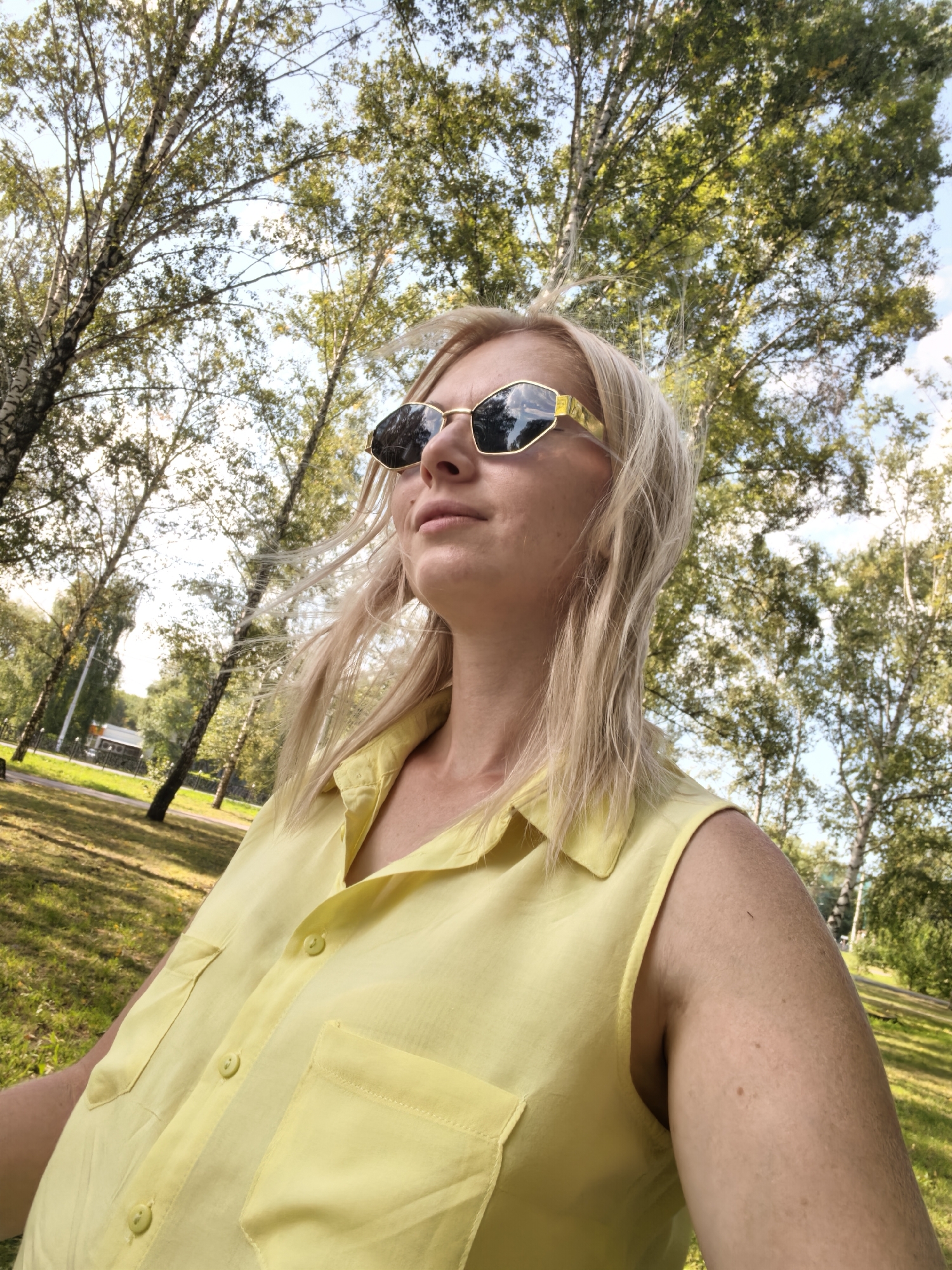 A blonde woman wearing a yellow sleeveless shirt and gold-rimmed sunglasses is shown in a medium close-up. She is looking upwards and to the left, with her hair blowing in the wind. The background features a park with trees and a clear sky, suggesting an outdoor setting. The image was taken in Sterlitamak, Russia.