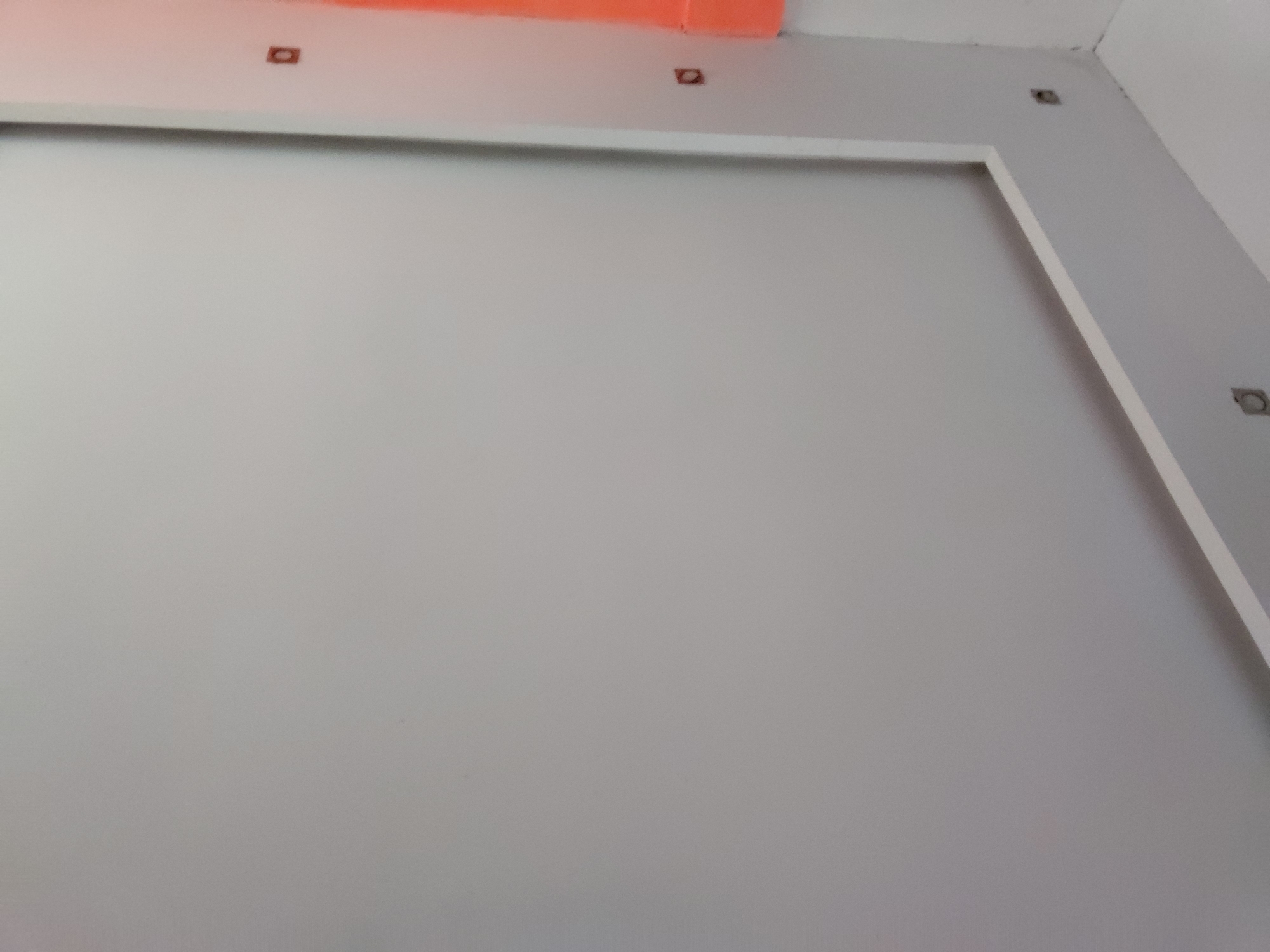 The image displays an interior view of a room, likely under construction or renovation, in Santa Ana, Venezuela. The dominant feature is a large, light grey wall, which appears to be freshly painted or primed. A prominent white trim or frame outlines a significant rectangular section on this grey wall, suggesting a decorative panel or a prepared area for a screen or artwork.
Above this framed section, towards the top of the wall and extending into the ceiling area, several small, dark, rectangular openings or cutouts are visible. These holes are evenly spaced and appear to be prepared points for electrical outlets, light fixtures, or mounting hardware.
The ceiling above the wall is partially visible. One section of the ceiling is painted a distinct orange or salmon color, running horizontally across the top of the frame. Adjacent to this, another part of the ceiling is white. In the upper right corner, where the grey wall meets another wall and the ceiling, some unfinished plastering or painting is evident, indicating ongoing work.
There are no people or specific activities depicted in the image. No text is visible. The lighting is uniform, suggesting indoor conditions without direct sunlight.