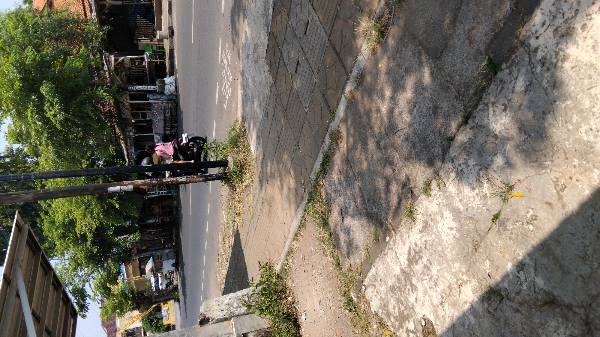The image depicts a street scene in Proyonangg, Indonesia, on a sunny day. A motorcycle carrying two individuals is moving on the asphalt road. The person in the rear is wearing a pink shirt and a helmet. On the side of the road, a footpath is visible, constructed with square tiles and concrete slabs. Scattered grass and weeds grow along the edges of the footpath and on the concrete. In the background, there are trees, small shops or stalls, and buildings. The road has white dashed lines indicating lanes. The overall environment suggests a typical urban or semi-urban setting with modest commercial activity.