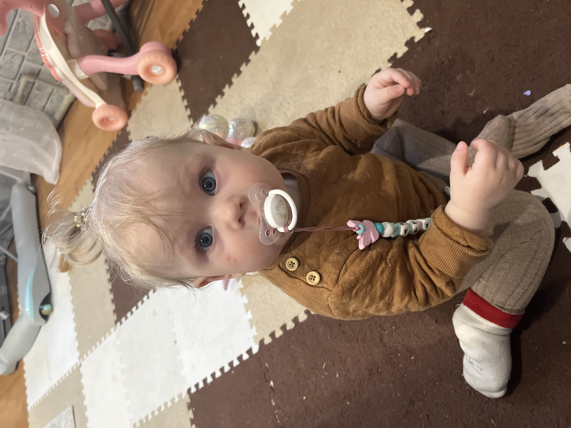 An overhead shot captures a baby with light blonde hair and blue eyes, sitting or lying on a foam puzzle mat. The baby is wearing a brown long-sleeved top with three visible wooden buttons, grey pants, and white socks with red cuffs. A pacifier, adorned with a colorful clip featuring pink, blue, and white beads and a pink butterfly charm, is in the baby's mouth. The baby gazes directly at the camera with a neutral yet curious expression, with a few small red spots visible on one cheek.
The setting is an indoor space, likely a home in Vesyoloye, Russia, identified by the interlocking brown, beige, and white foam puzzle mat pieces covering the floor. In the background, to the baby's left, a pink child's ride-on toy with wheels is partially visible. Near the baby's left arm, several iridescent or glittery round objects are scattered on the mat. A white object, possibly a piece of furniture or another toy, is also partially seen on the far left edge of the frame. The floor appears to have some dust or small debris. No visible text is present.