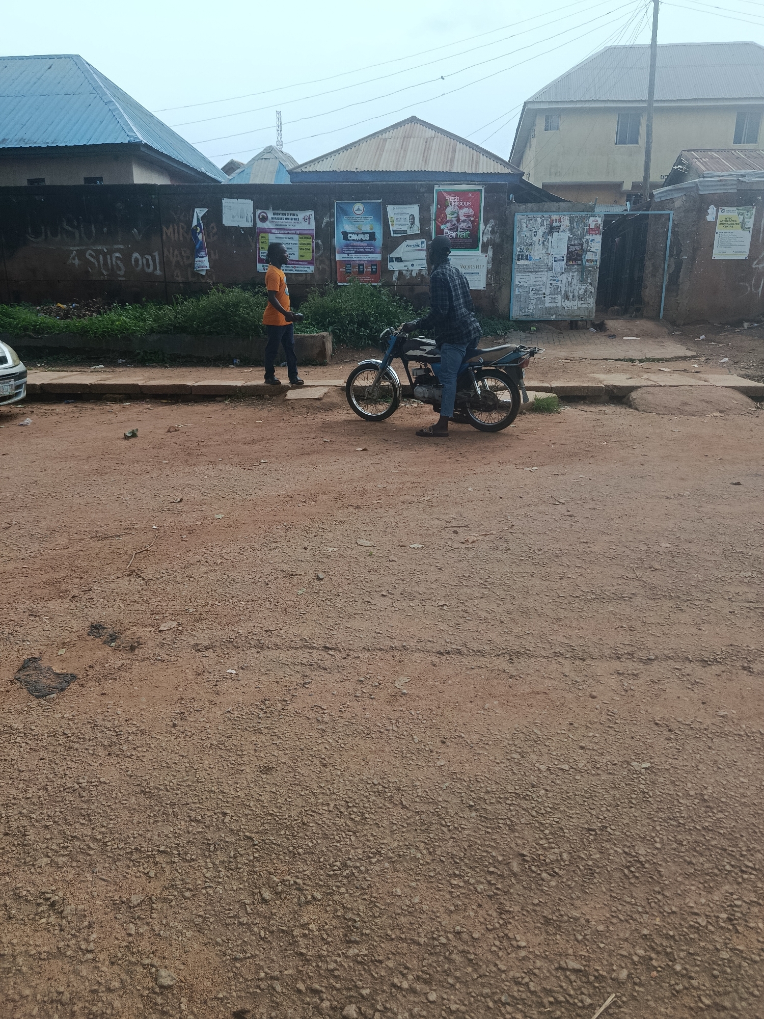 In Rafin Par, Nigeria, a dirt road in a residential area is depicted. The road is dusty and uneven, with scattered debris and small stones. On the left side of the road, a white car is partially visible. In the background, a wall is adorned with posters and graffiti. To the right of the wall, a man wearing an orange t-shirt and dark pants stands on the sidewalk. Further to the right, another man dressed in a plaid shirt and blue jeans is seated on a motorcycle, with the motorcycle parked on the road. Buildings with corrugated metal roofs are visible in the background, along with utility poles and wires. The sky is overcast, suggesting a cloudy day.