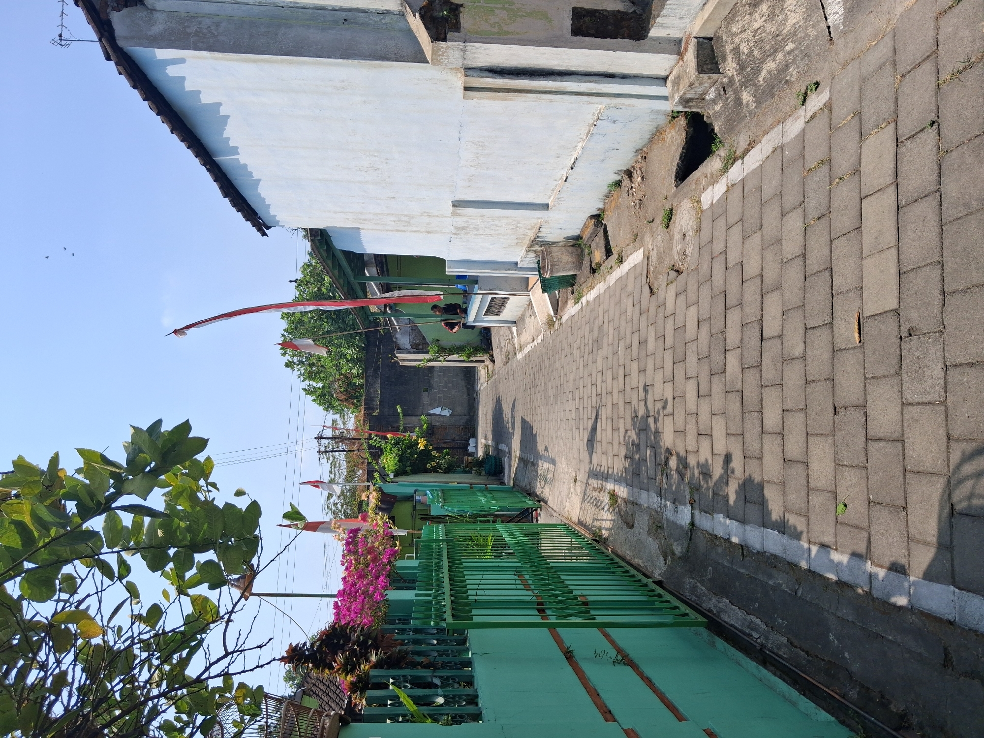 This is a ground-level, full-shot photograph of a narrow, paved alleyway in Surakarta, Indonesia. The alleyway is lined with houses and fences. The paving stones are a light grey color, and the alley is illuminated by bright sunlight, casting shadows. On the left side of the alley, there is a bright green metal fence with intricate detailing. Behind the fence, there are lush green trees and vibrant pink flowers. Two Indonesian flags, red and white, are visible on poles in the background. On the right side of the alley, a light blue-colored house with a dark tiled roof stands prominently. The wall of the house appears to be weathered and slightly stained. In front of the house, there are concrete structures and a trash can. Two individuals are partially visible on the porch of the house on the right, appearing to be interacting. The overall atmosphere suggests a quiet, residential street.