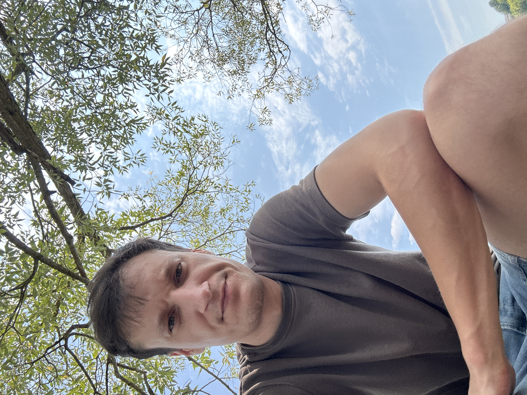 A low-angle, close-up, selfie-style photo captures a man from the chest up, positioned outdoors. The man, with fair skin and short brown hair, wears a dark grey, textured t-shirt and denim shorts. He is seated, with his left arm resting on his crossed legs and his right arm bent at the elbow. His gaze is directed at the camera with a neutral expression. The background is dominated by the sky, partially obscured by the branches and leaves of a deciduous tree. The sky is a clear blue with scattered white clouds. The overall setting suggests a park or outdoor recreational area within the city of Makiv, Ukraine.