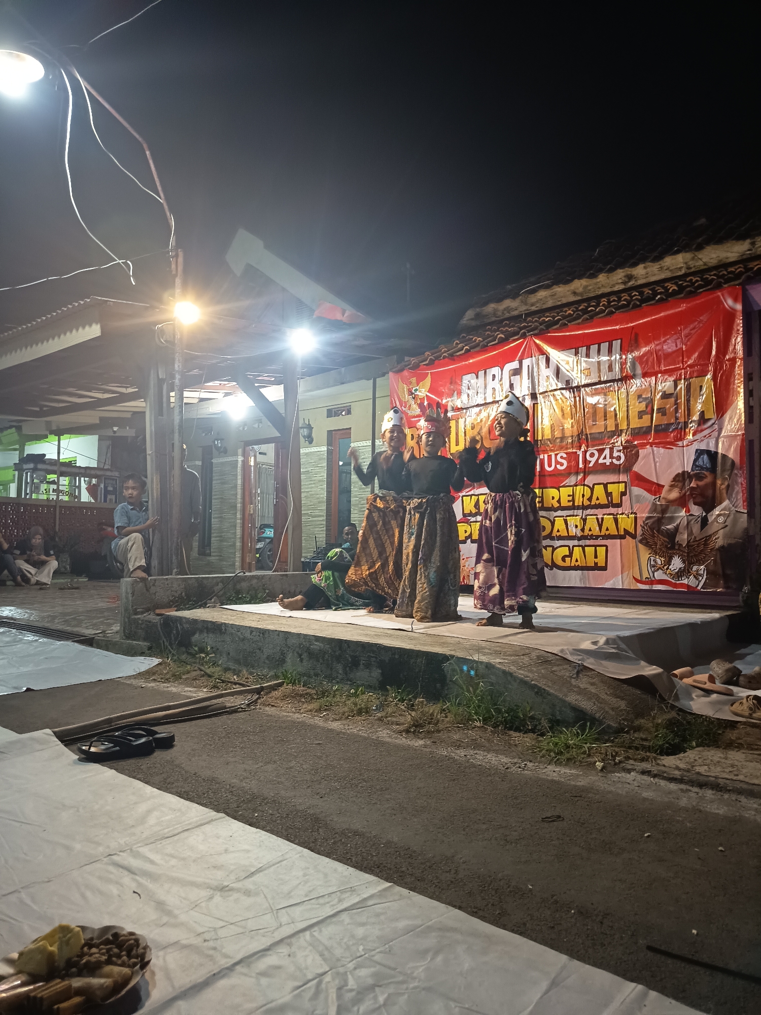 A group of four children dressed in traditional attire are performing on a stage at night. A red banner with Indonesian text and an image of a salute is hung behind them. The stage is set up outdoors, with a few onlookers seated on the ground. The scene appears to be in a residential area, with houses and trees visible in the background.