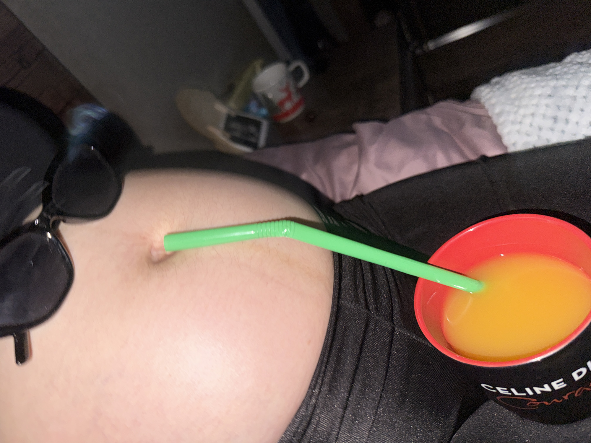 The image shows a close-up view of a person's belly with a green straw inserted into their belly button. The person is wearing dark clothing. To the right of the belly is a red cup filled with an orange liquid, presumably a beverage. The cup has text on it that appears to be "CELINE D..." and possibly "Courage" or a similar word. In the background, out of focus, there are other objects including a white mug with red designs, a pair of light-colored shoes, and a pink fabric. The lighting suggests it might be evening or indoors. The overall scene is casual and potentially humorous, with the straw in the belly button being the focal point. There are no clear indicators of the weather or the specific location beyond the provided context of Tbilisi, Georgia. The image focuses on the unusual placement of the straw, creating a quirky and attention-grabbing composition.