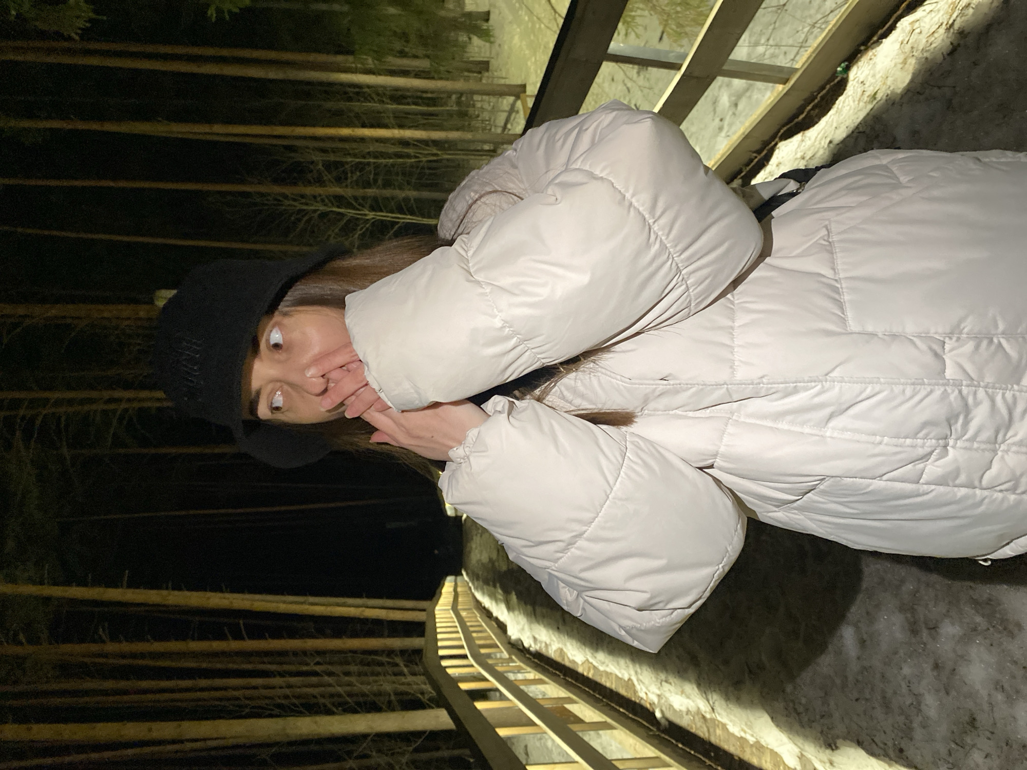 A person wearing a black beanie and a light-colored puffer jacket stands on a wooden walkway in an outdoor setting that appears to be a park or wooded area at night. The person is covering their mouth with their hand and looking to the side with a surprised expression. The walkway has wooden railings and leads down a slope. Tall trees are visible in the background, illuminated by artificial light. The location of the image could be in Votkinsk, Russia, or another place, this can not be determined with the information available.