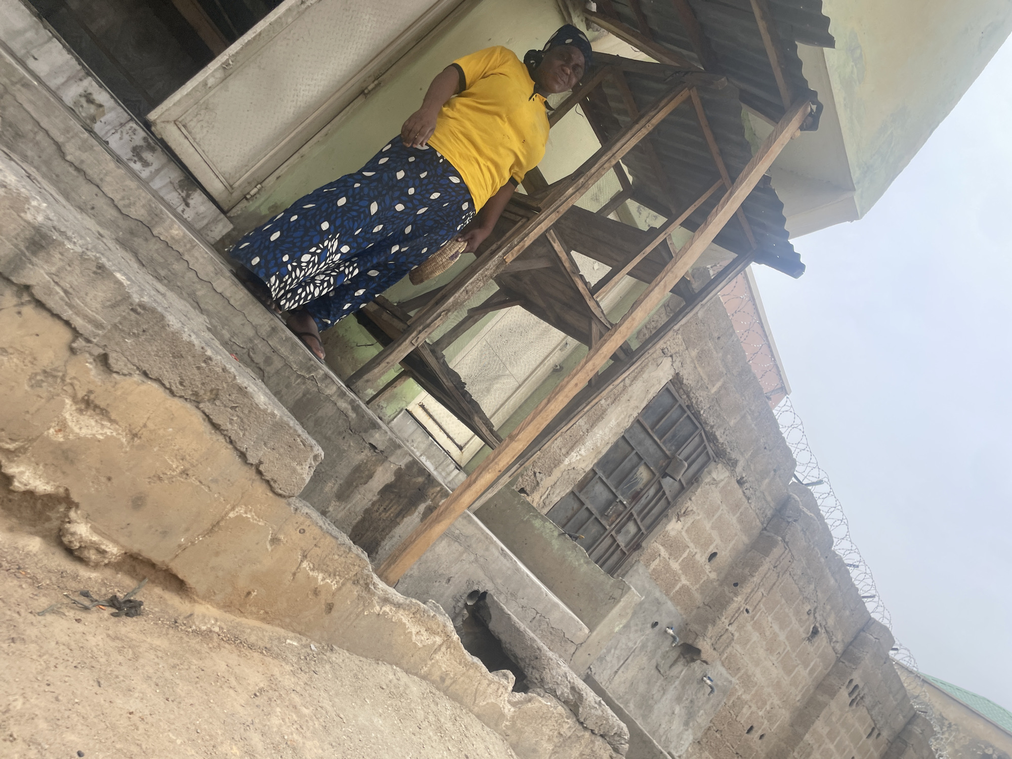 An eye-level, medium shot presents a woman in a yellow top and a blue, patterned skirt standing on a concrete structure adjacent to a partially collapsed or under-construction building. The woman appears to be holding a woven object. The building features exposed wooden beams and incomplete corrugated metal roofing. The environment is urban, with a view extending to an overcast sky. The image is slightly rotated, and the location is identified as Unknown, Unknown. A detail of the building shows a brick-like construction with barbed wire along the top.