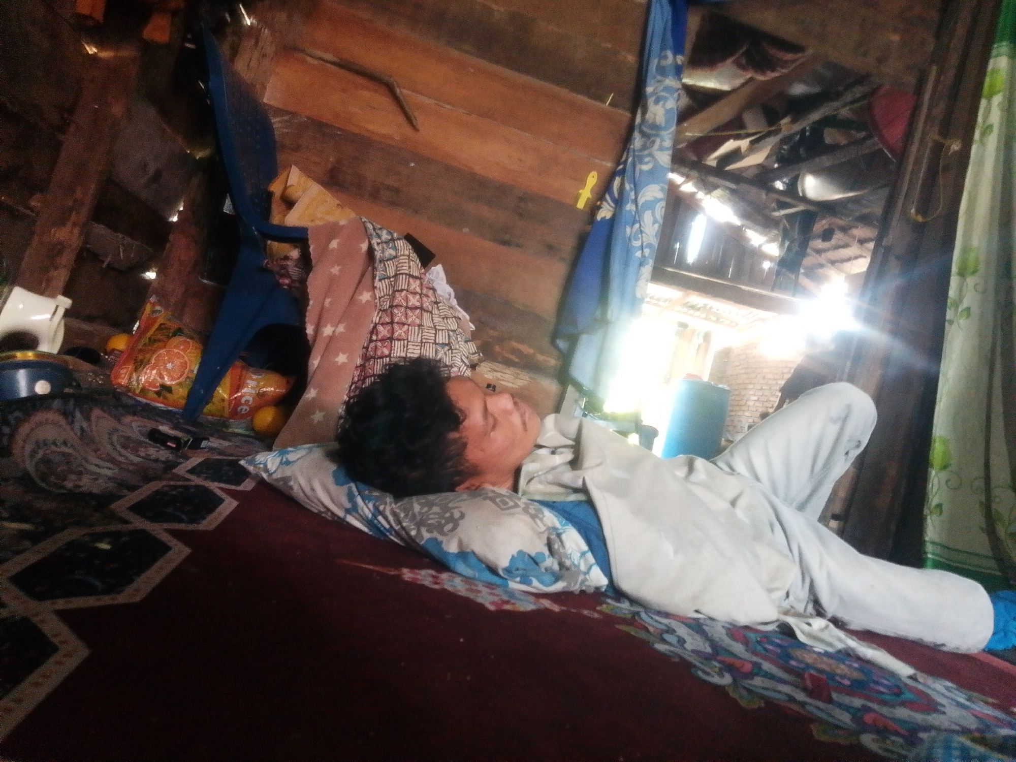 A young person with dark, curly hair lies on their back, eyes closed, on a pillow on a red carpeted floor within a rustic wooden structure. The person is covered by a light-colored garment, possibly a shirt and pants. The room appears to be dimly lit, with sunlight streaming in from the right side of the frame. Various objects are visible in the background, including what appears to be a blue chair, colorful fabric draped on the wall, and packaging for food items. The floor is covered with a patterned carpet. The environment suggests a simple, possibly rural dwelling in Labuhan Bilik, Indonesia.