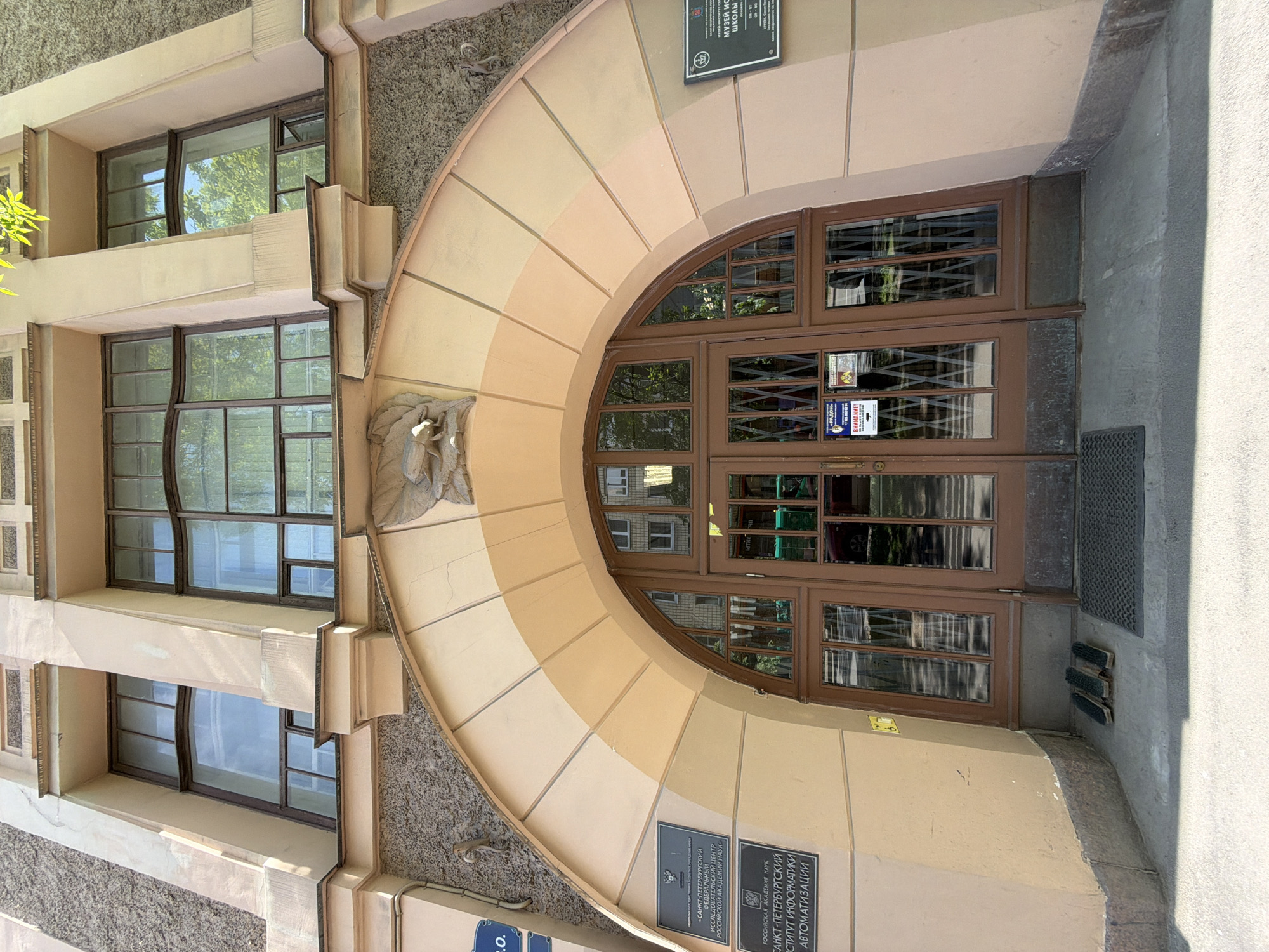 The image displays the entrance of a building in Saint Petersburg, Russia, characterized by a large, semicircular archway that frames double wooden doors. The archway is constructed from segmented blocks, transitioning in color from lighter cream to darker beige. Beneath the arch, to the left of the doors, there is a carved decorative relief, possibly depicting a bird or mythical creature.
The building's facade combines smooth, pale beige plaster with areas of textured, rough grey material. On the left side, multiple windows with dark brown frames and intricate glass panes are visible, some exhibiting a slight distortion. Reflections in the glass show lush green trees and parts of the surrounding environment, suggesting a clear, sunny daytime.
The tall entrance doors are made of dark wood and feature numerous small, gridded glass panes. Various stickers are affixed to the doors, including a red one with "ВНИМАНИЕ!" (Attention!). A dark doormat and shoe brushes are present at the base of the doors.
Several informational plaques are mounted on the building. On the lower right, two plaques identify the location: "РОССИЙСКАЯ АКАДЕМИЯ НАУК" (Russian Academy of Sciences) and "САНКТ-ПЕТЕРБУРГСКИЙ ИНСТИТУТ ИНФОРМАТИКИ АВТОМАТИЗАЦИИ" (Saint Petersburg Institute for Informatics and Automation). Another plaque, partially visible above the arch, reads "МУЗЕЙ ИСТОРИИ ШКОЛЫ" (Museum of School History). The full text for the institute also includes "САНКТ-ПЕТЕРБУРГСКИЙ ФЕДЕРАЛЬНЫЙ ИССЛЕДОВАТЕЛЬСКИЙ ЦЕНТР РОССИЙСКОЙ АКАДЕМИИ НАУК" (Saint Petersburg Federal Research Center of the Russian Academy of Sciences). No people are visible, and no specific activity is taking place, presenting a static view of the building's exterior.