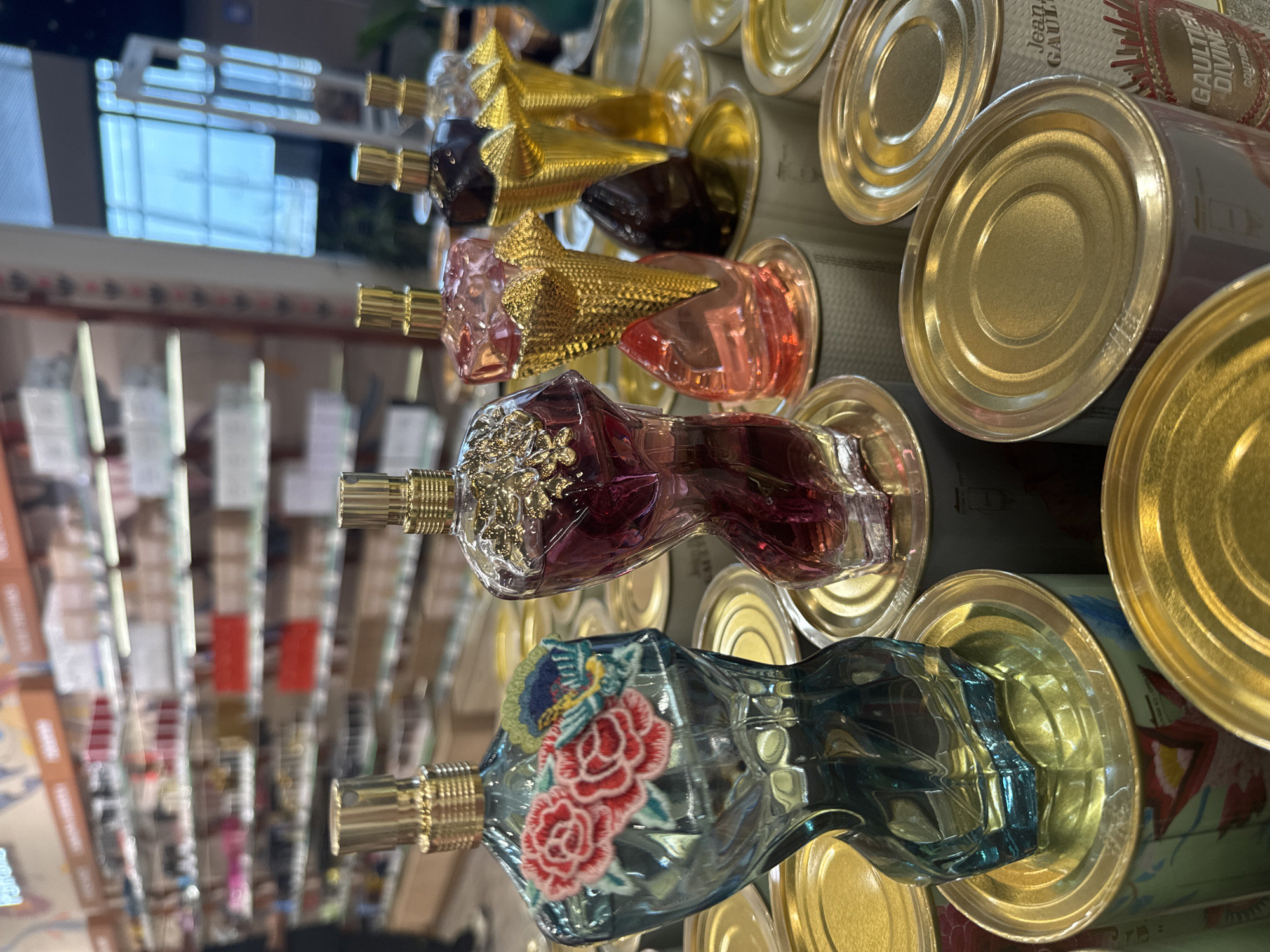 The image depicts a display of Jean Paul Gaultier perfume bottles arranged on top of cylindrical cans. The bottles, varying in color, are shaped like a woman's torso and feature ornate, gold-colored caps. The cans supporting the bottles appear to be part of the packaging or display itself. The overall setting is likely a retail environment, possibly a department store or perfume shop. The location is suggested to be Tashkent, Uzbekistan, though there are no explicit identifiers in the image itself to confirm this.