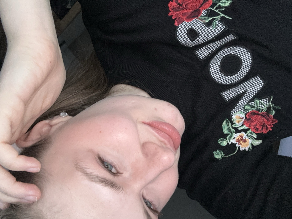 A woman lies on her side, facing the camera, with a relaxed expression. She wears a black sweater with the word "NOIR" embroidered on it. The letters are decorated with a houndstooth pattern and accompanied by red rose designs. Her hair is light brown, and she wears a small earring. Her skin tone is fair. Her fingers are near her head.